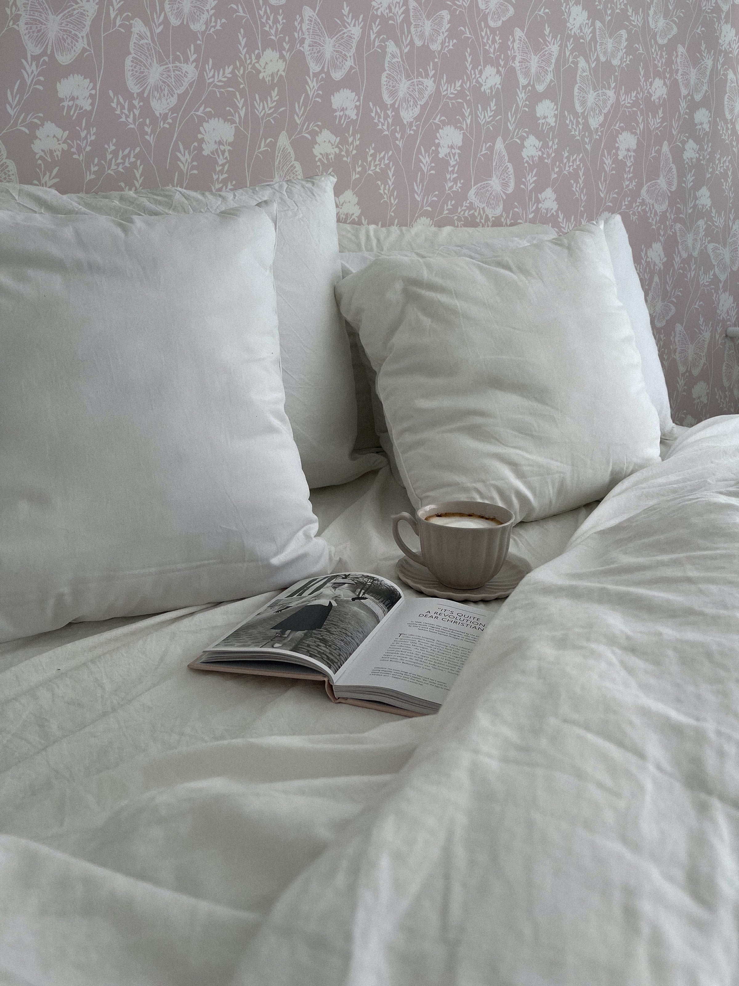 a cozy bedroom setting where the "Whimsical Wings Wallpaper" provides a whimsical background. The camera captures a part of the bed with plush pillows, a book, and a cup of coffee, inviting a relaxed and homey feel complemented by the soft wallpaper design.