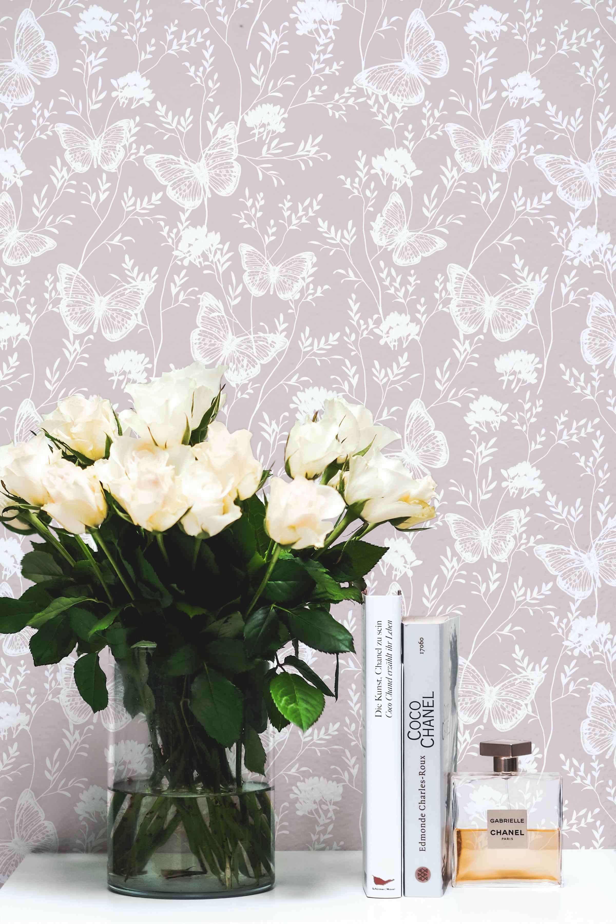 a closer look at the "Whimsical Wings Wallpaper," where a bouquet of cream roses in a clear vase is positioned against the backdrop. The delicate butterflies and floral outlines on the wallpaper add a sophisticated and gentle touch to the decor.
