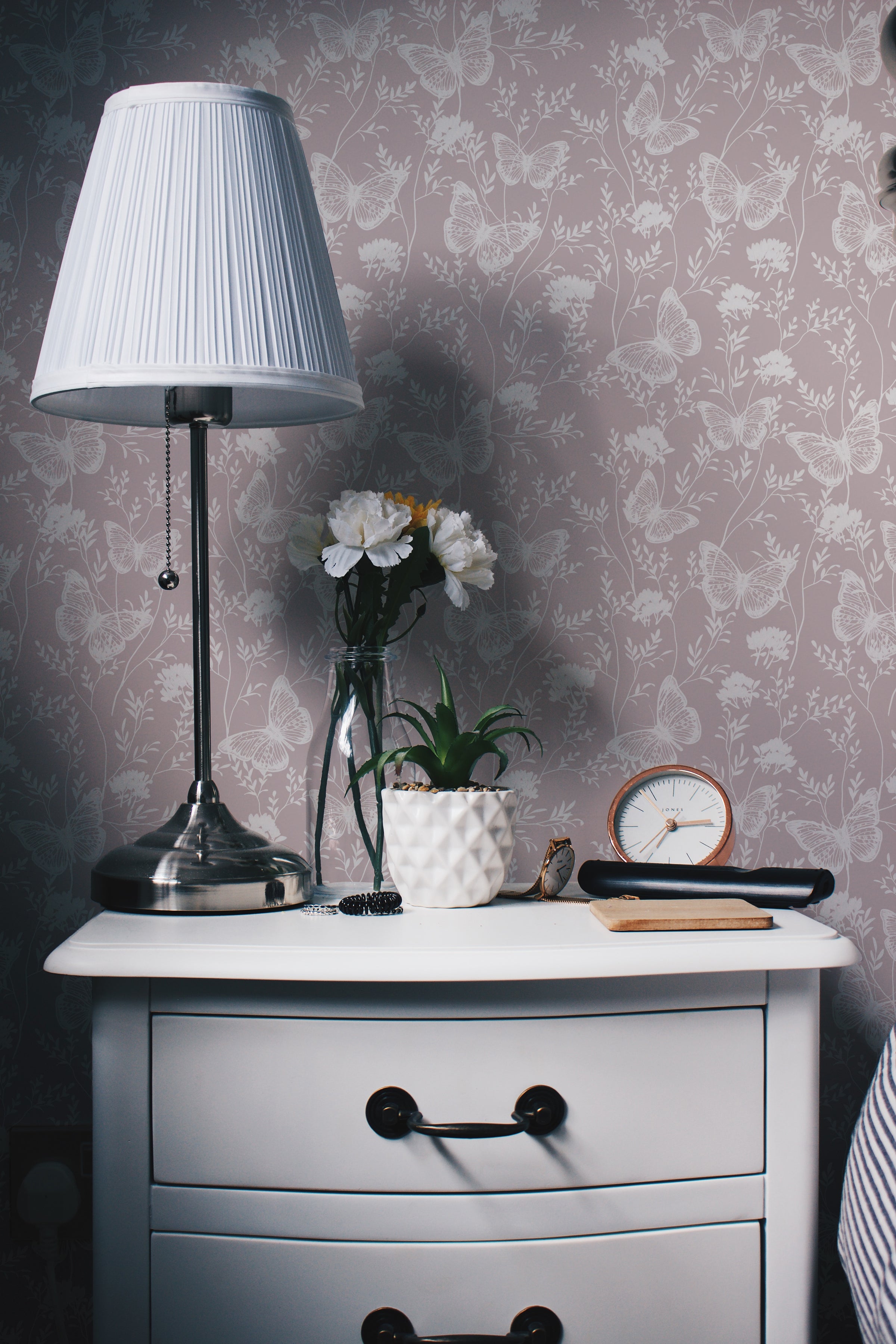 an image of a bedroom corner with the "Whimsical Wings Wallpaper" adorning the wall. The wallpaper features a soft mauve-pink background with an elegant pattern of white butterflies and floral elements, creating a tranquil and romantic atmosphere. A classic silver lamp and white flowers on a nightstand contribute to the room's serene ambiance.
