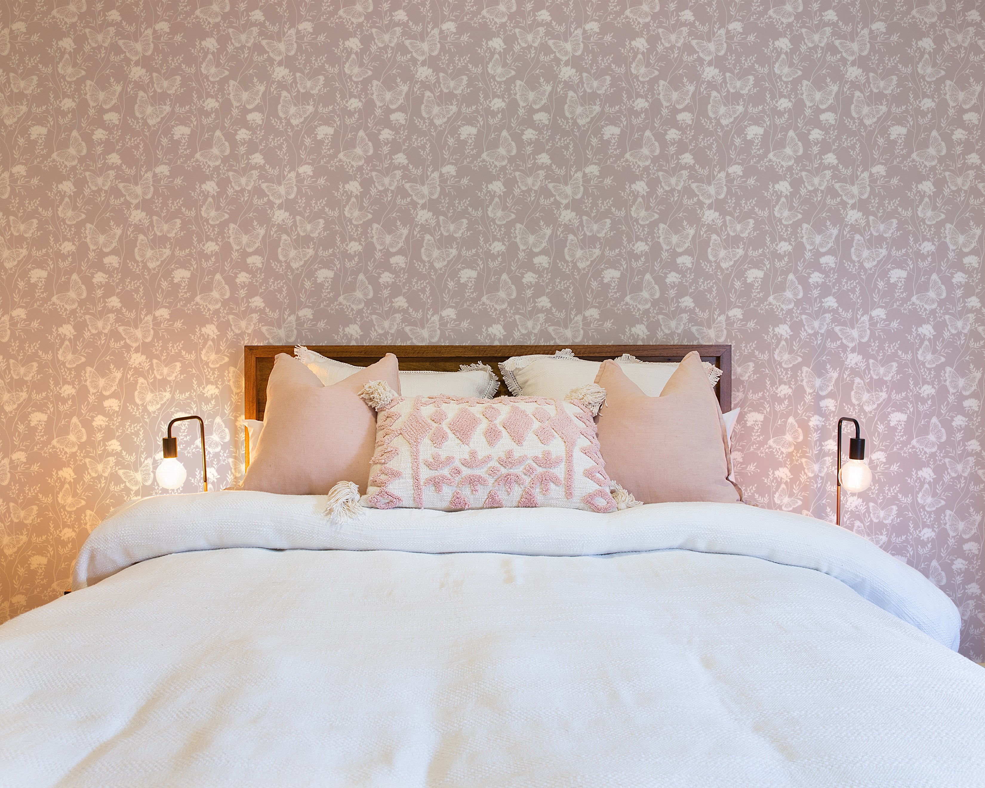 the "Whimsical Wings Wallpaper" in a warmly lit bedroom. A stylish bedside table with a minimalist lamp showcases the gentle butterfly pattern on the wallpaper, pairing beautifully with the room's neutral and pastel color scheme for a peaceful sleeping space.