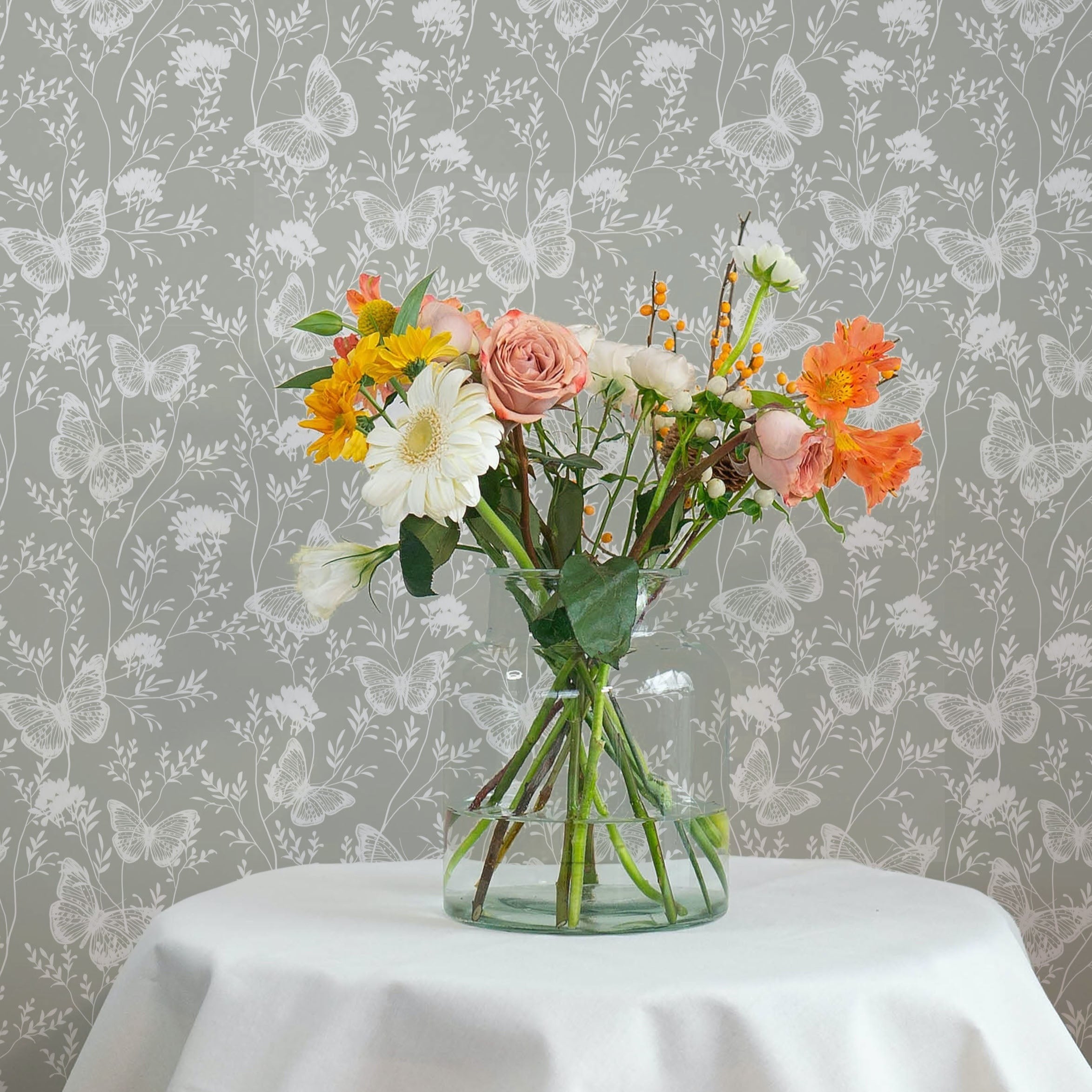 An elegant home decor arrangement using the Whimsical Wings Wallpaper-Olive, with a vase of vibrant, colorful flowers set against the wallpaper, highlighting its delicate butterfly and floral motifs.