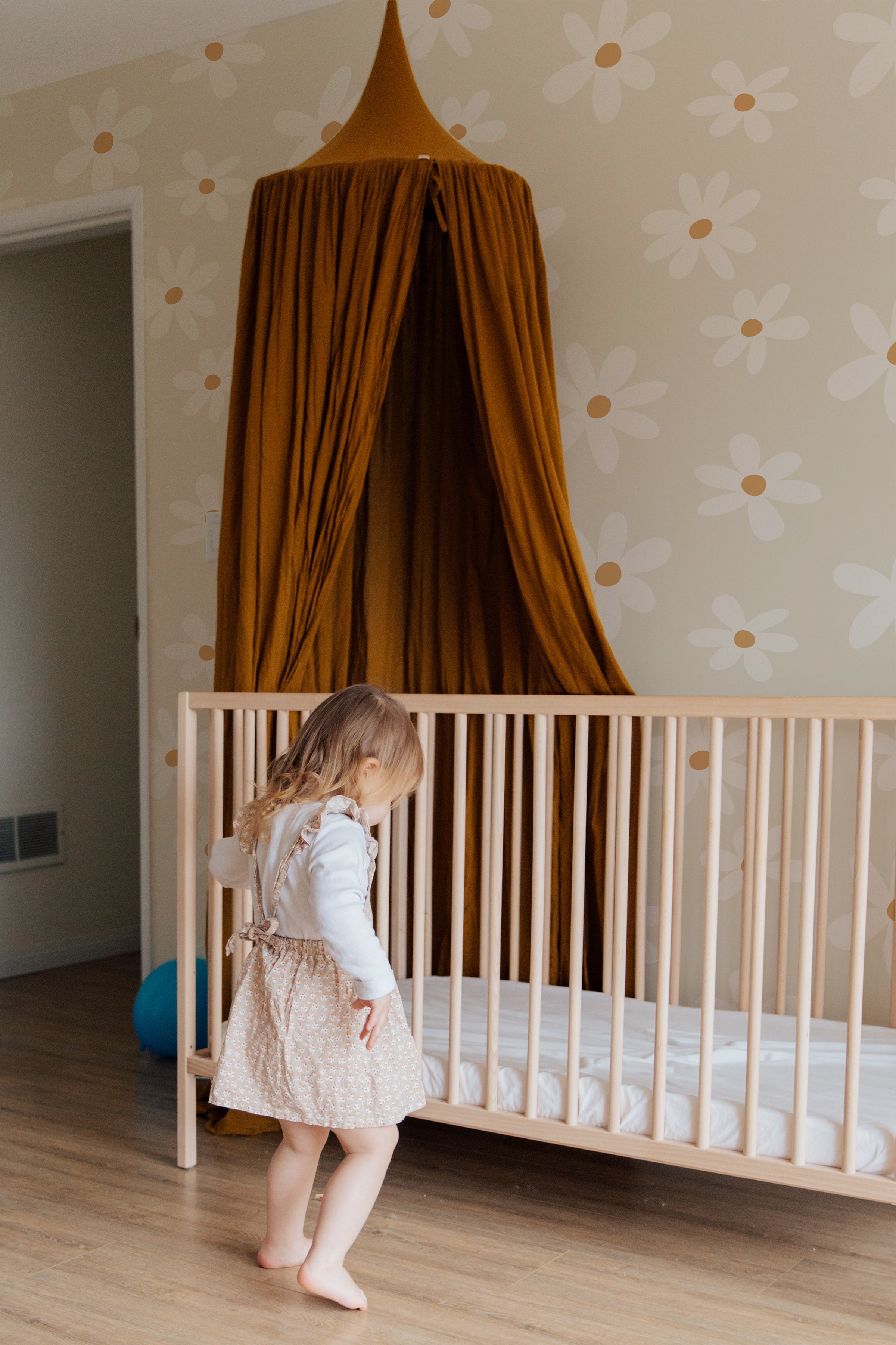 A child's nursery room with a wall adorned with Simple Daisy Wallpaper, featuring large white daisies with golden centers on a soft ecru background. A little girl in a white blouse and patterned skirt stands beside a wooden crib, beneath a mustard-colored canopy.