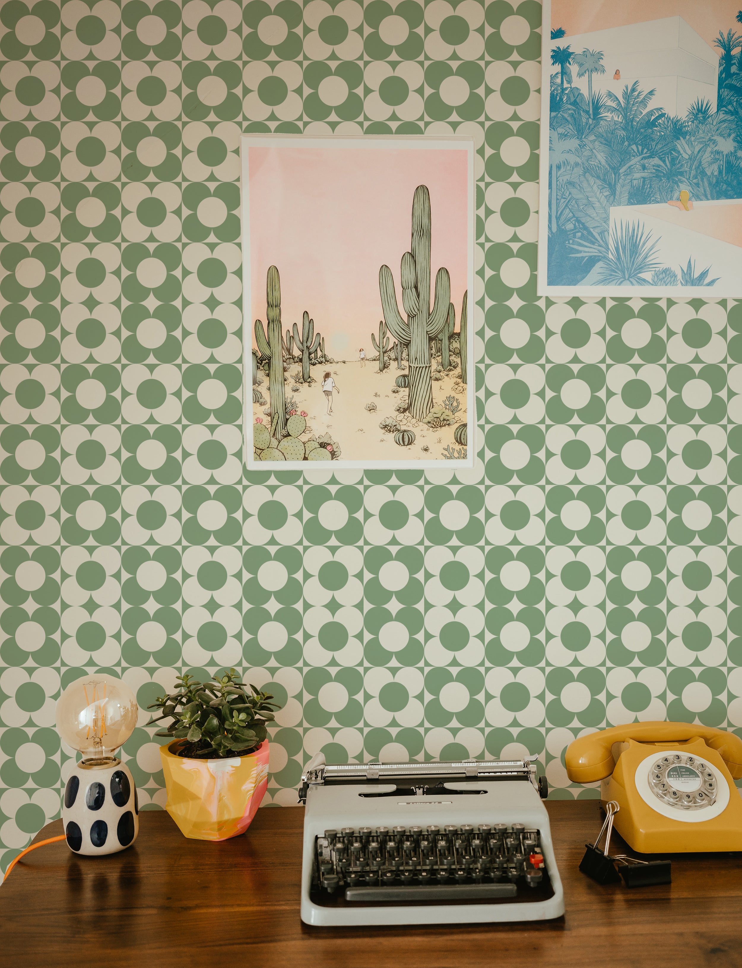 A retro-styled home office space featuring the Vintage Groovy Wallpaper - Green. The wall is covered with a green and white circular geometric pattern that complements the vintage decor including a yellow rotary phone, a classic typewriter, and colorful desk accessories, creating a cheerful and nostalgic atmosphere