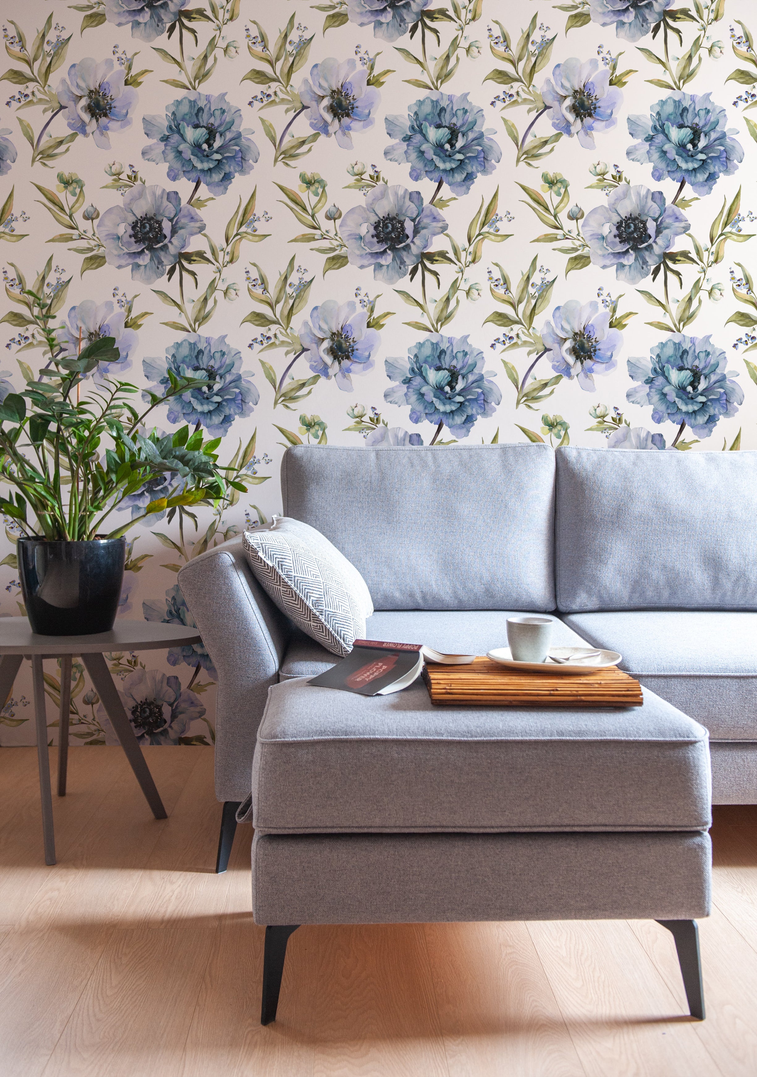 A stylish living room showcasing Indigo Floral Wallpaper, with large, beautifully detailed blue flowers against a soft white background, complementing the light grey fabric sofa and enhancing the room’s elegant aesthetic.