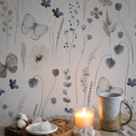 Romantic and warm atmosphere highlighted by Soft Flutter Wallpaper in a dimly lit setting with candles. The wallpaper's soft botanical patterns and butterflies provide a tranquil backdrop to a cozy setting with a mug and candles on a wooden tray