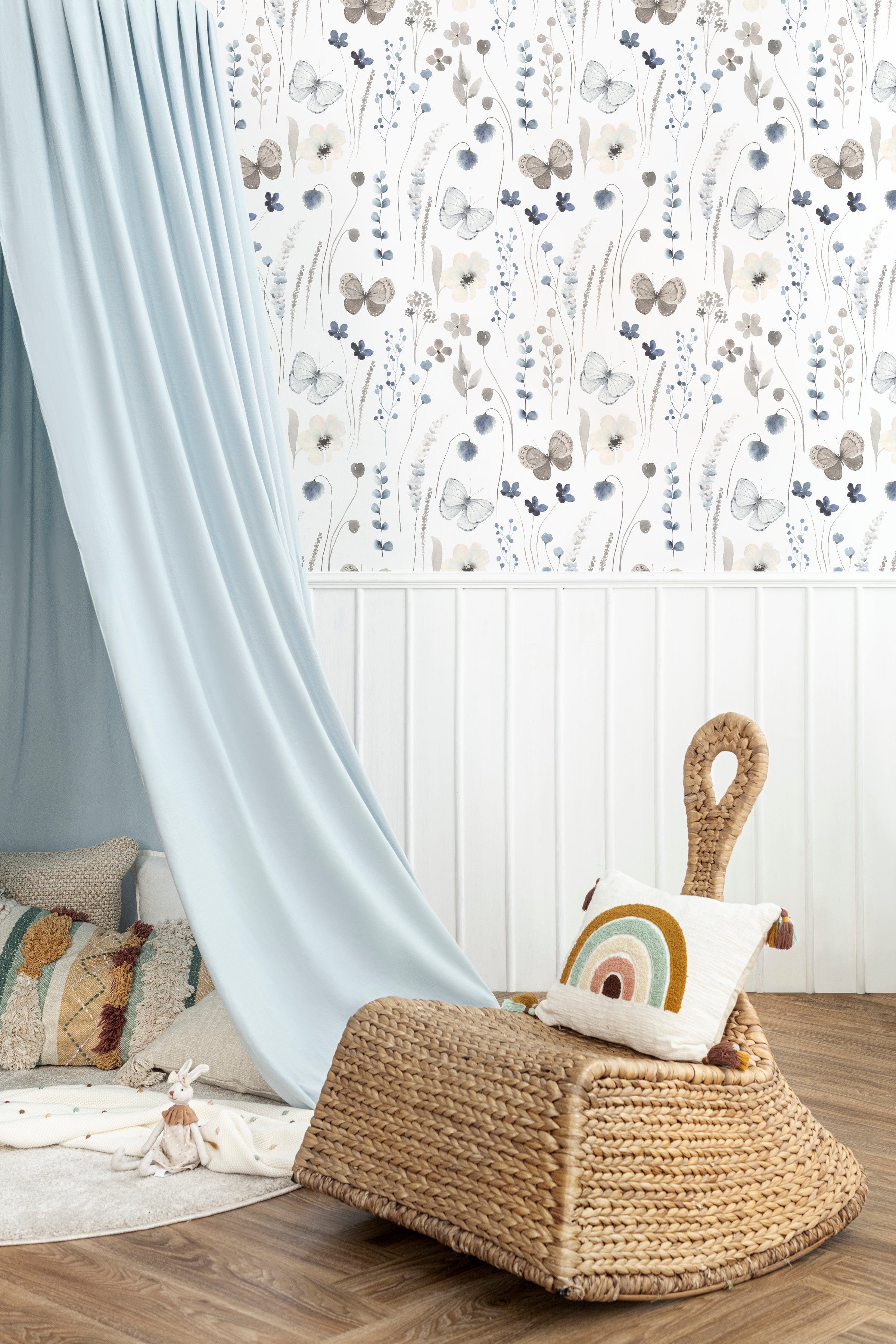 Cozy nook in a room adorned with Soft Flutter Wallpaper, which displays an elegant botanical and butterfly pattern in soft blues and grays. A light blue curtain partly reveals a textured wicker lounge chair with colorful pillows, situated against a half paneled white and wallpapered wall