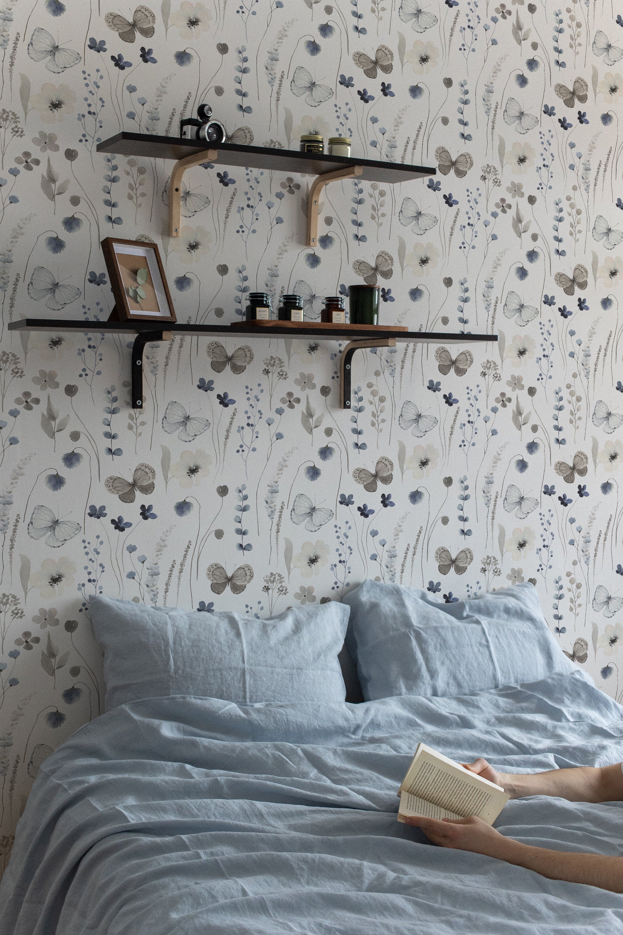 Bedroom scene featuring Soft Flutter Wallpaper with a serene butterfly and floral motif in muted colors. A bed with soft blue linen is partially visible, enhancing the peaceful ambiance, complemented by wooden shelves with minimalist decor