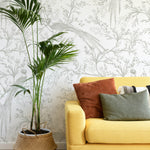 A chic living room showcasing Oriental Garden Wallpaper, featuring a subtle and elegant botanical design with birds perched on branches. The soft gray and white color palette creates a tranquil backdrop for the vibrant yellow sofa and a large indoor palm plant in a woven basket, enhancing the room's natural elegance.