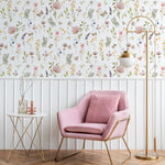 A modern interior featuring the Floral Fields Wallpaper, with its delicate array of wildflowers and butterflies in soft pinks, purples, and yellows, creating a fresh and serene ambiance. A plush pink armchair with gold accents, a white side table, and a stylish gold floor lamp complement the wall's floral design.