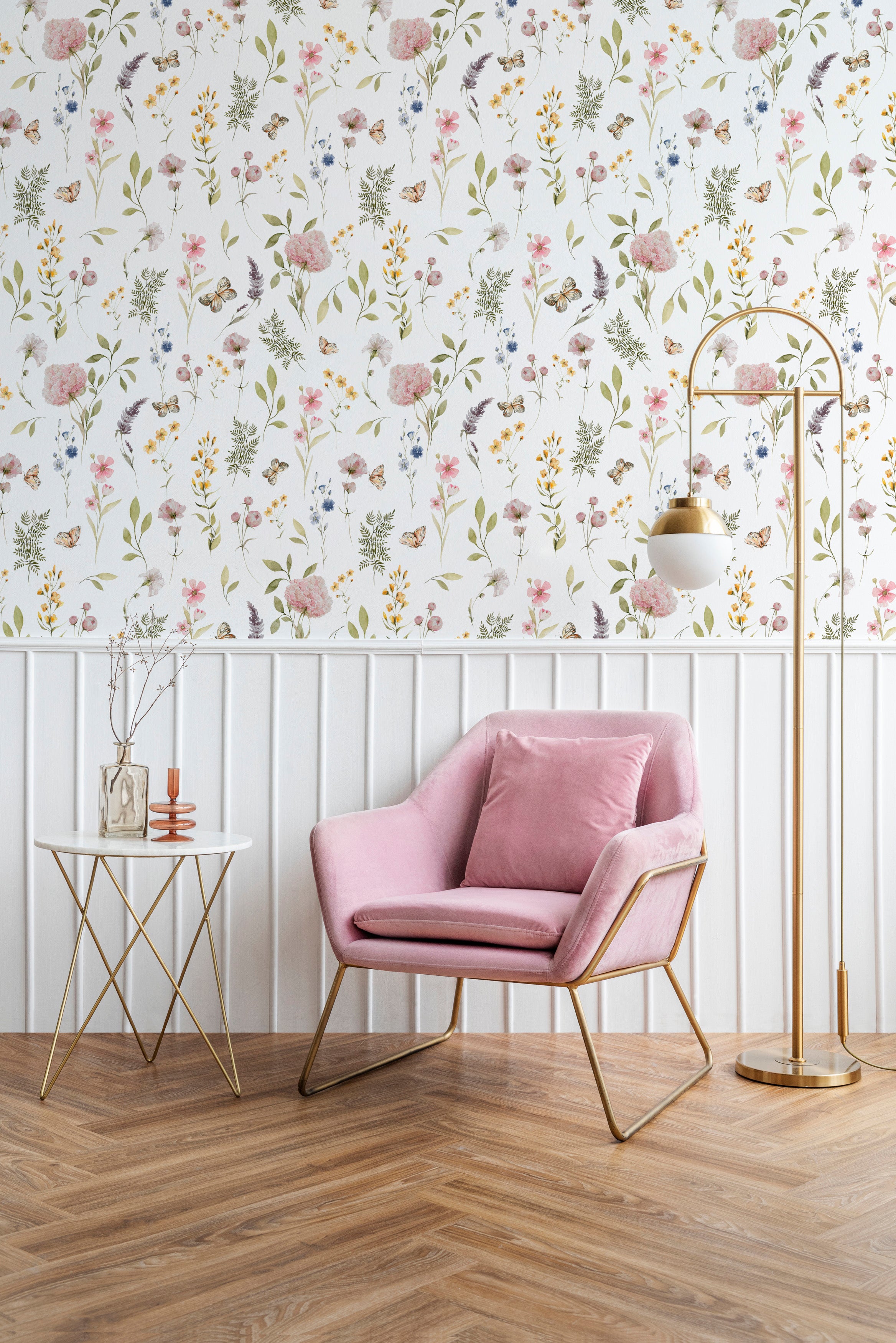 A modern interior featuring the Floral Fields Wallpaper, with its delicate array of wildflowers and butterflies in soft pinks, purples, and yellows, creating a fresh and serene ambiance. A plush pink armchair with gold accents, a white side table, and a stylish gold floor lamp complement the wall's floral design.
