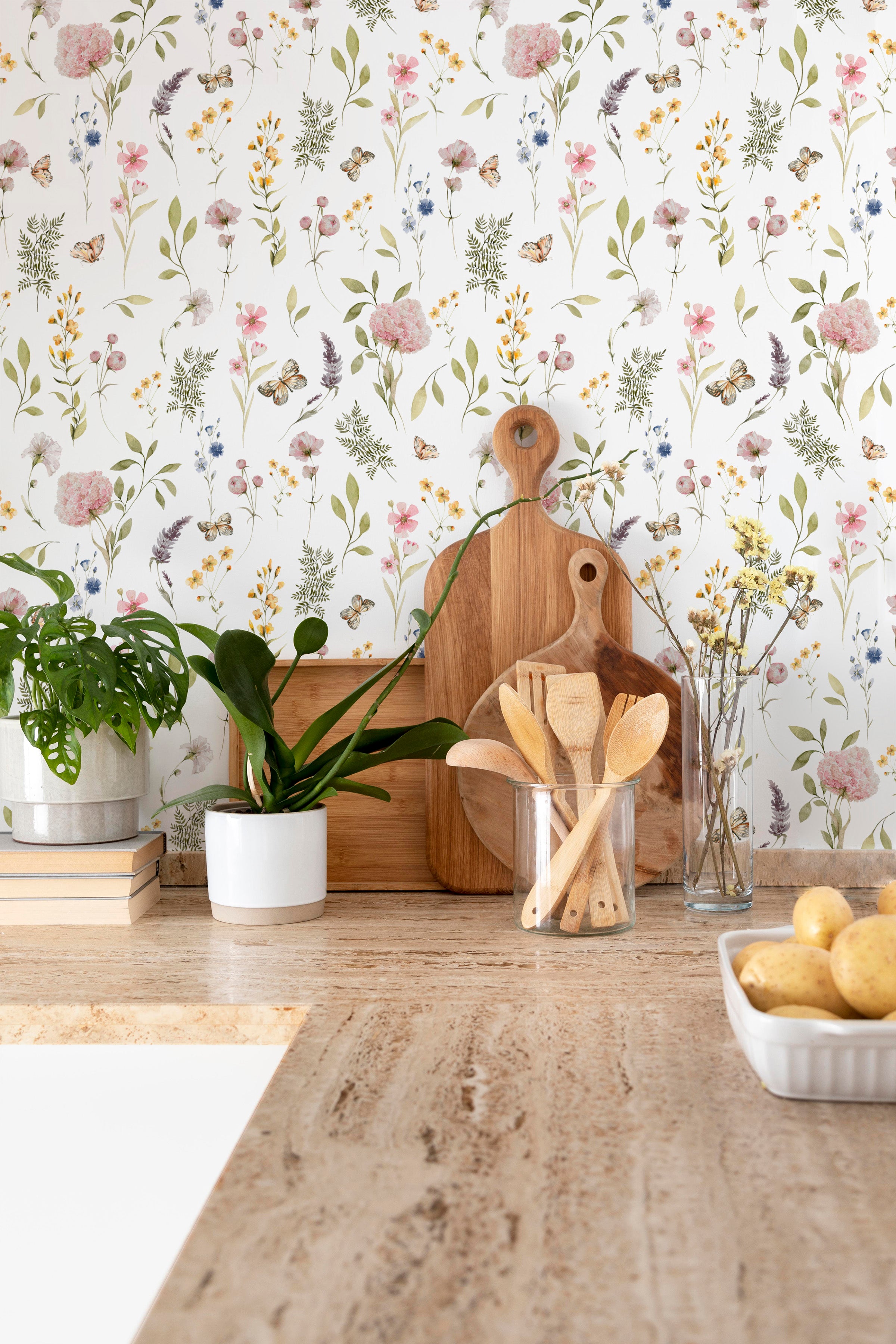 A well-lit kitchen corner showcases the Floral Fields Wallpaper as a backdrop, enhancing the space with its soft floral print. Wooden kitchen accessories, fresh plants, and a clear vase of dried flowers sit atop a countertop, harmonizing with the wallpaper's botanical theme.