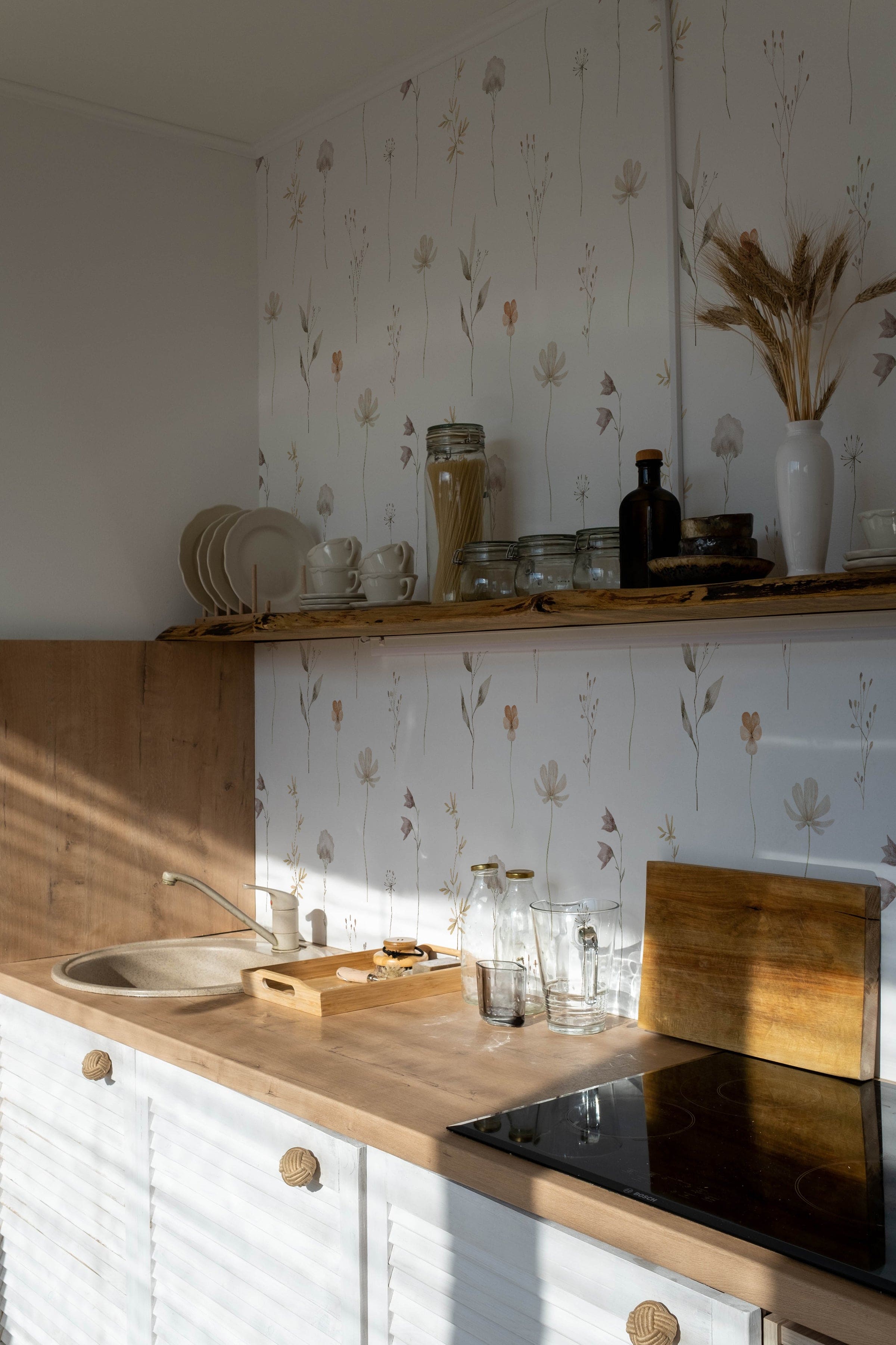 A kitchen corner showcasing the 'Muted Floral Wallpaper' as a delicate backdrop behind open wooden shelves, where the simplicity of the floral designs complements the rustic charm of the kitchenware and natural wood textures