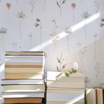 A serene composition of stacked books bathed in natural sunlight against a backdrop of 'Muted Floral Wallpaper', featuring delicate hand-drawn flowers and sprigs in soft neutrals that cast gentle shadows on the wall.