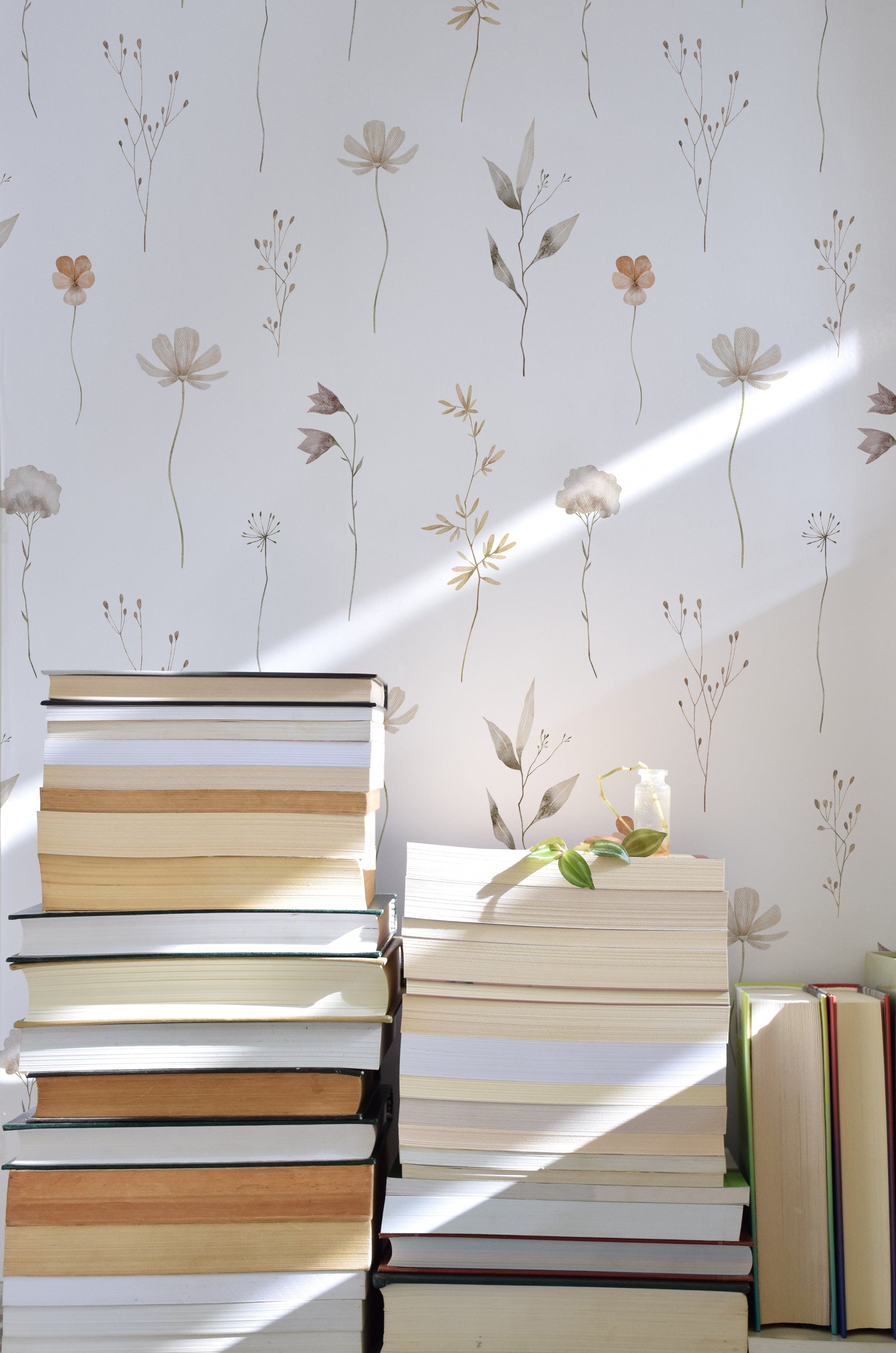 A serene composition of stacked books bathed in natural sunlight against a backdrop of 'Muted Floral Wallpaper', featuring delicate hand-drawn flowers and sprigs in soft neutrals that cast gentle shadows on the wall.