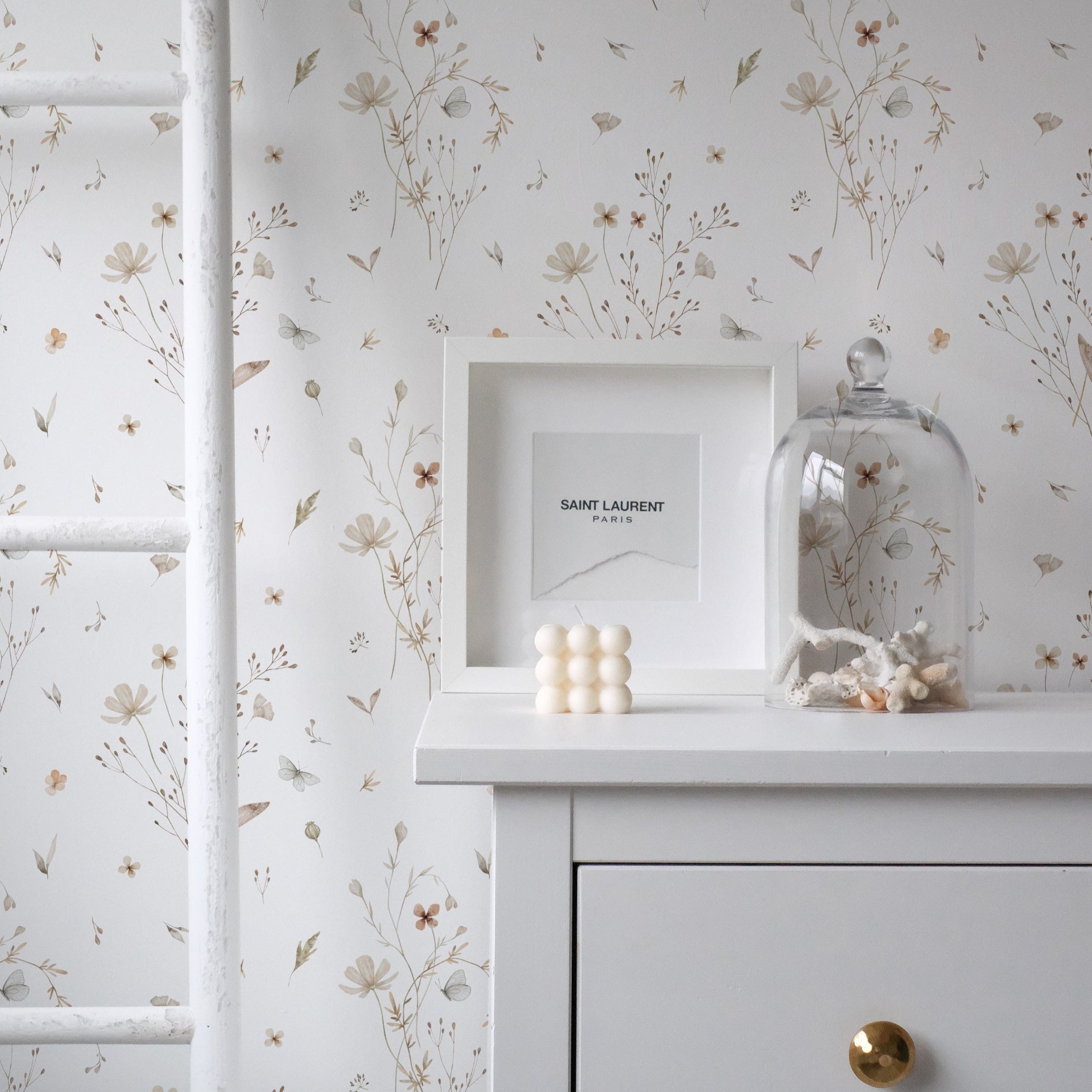 A close-up of a dresser against the Tranquil Bloom Wallpaper II, which is adorned with subtle illustrations of wildflowers and leaves in muted hues. The scene includes a white frame with a Saint Laurent logo, a bell jar with a collection of shells, and a cluster of decorative balls, creating a sophisticated and peaceful corner.