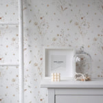  close-up of a dresser against the Tranquil Bloom Wallpaper II, which is adorned with subtle illustrations of wildflowers and leaves in muted hues. The scene includes a white frame with a Saint Laurent logo, a bell jar with a collection of shells, and a cluster of decorative balls, creating a sophisticated and peaceful corner.