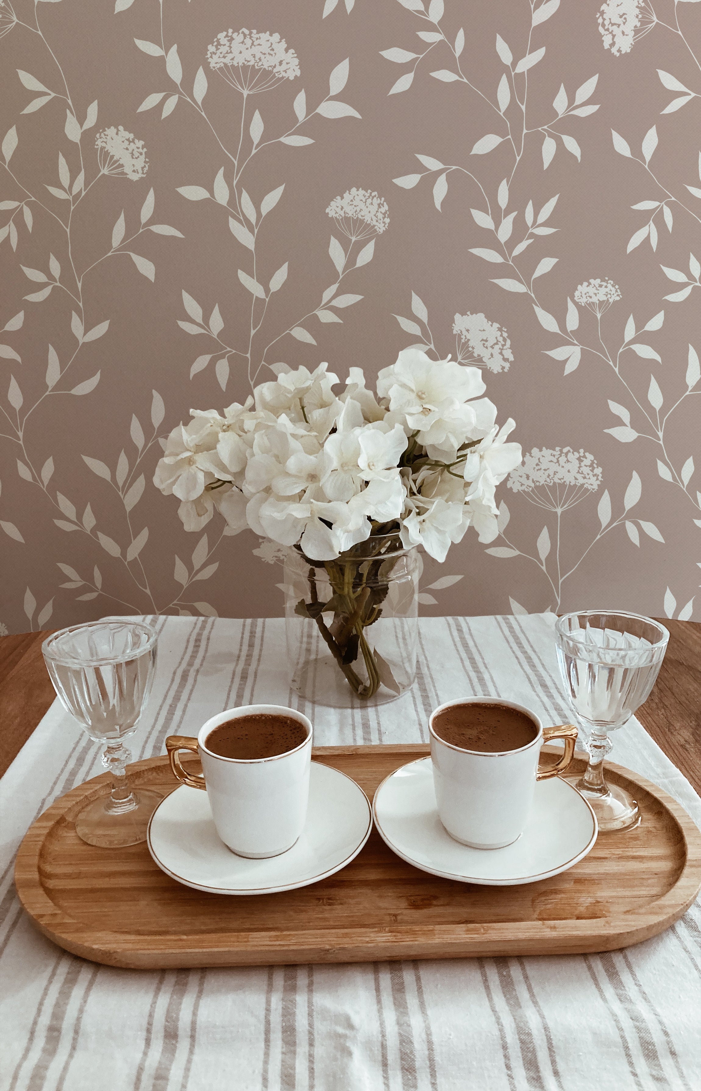 A cozy dining scene with a floral wallpaper in muted taupe, featuring elegant white flowers and delicate green foliage. A wooden tray on a table holds two cups of coffee and a vase with white blooms, enhancing the room's serene and inviting atmosphere.
