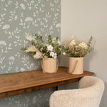  An interior setting demonstrating the application of the Botanical Elegance Wallpaper-Olive. The wallpaper provides a subtle yet charming backdrop for a wooden console table holding two cylindrical wooden vases with dried floral arrangements. The textures and colors of the foliage in the vases complement the wallpaper's botanical theme. To the right, the wall transitions to a solid olive shade, showing the wallpaper's potential for creating a cohesive color scheme in home decor.