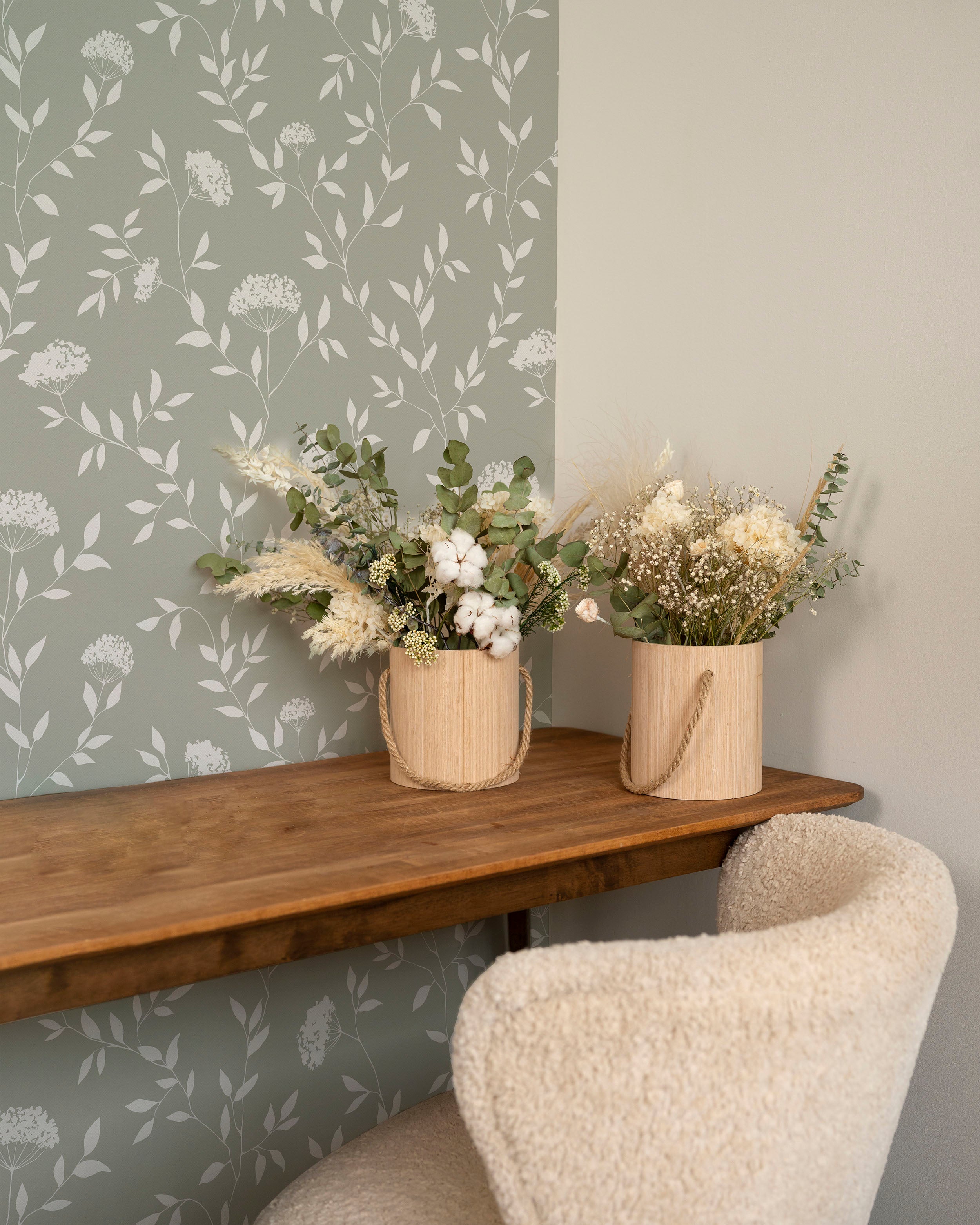  An interior setting demonstrating the application of the Botanical Elegance Wallpaper-Olive. The wallpaper provides a subtle yet charming backdrop for a wooden console table holding two cylindrical wooden vases with dried floral arrangements. The textures and colors of the foliage in the vases complement the wallpaper's botanical theme. To the right, the wall transitions to a solid olive shade, showing the wallpaper's potential for creating a cohesive color scheme in home decor.
