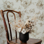 An elegant vintage wooden chair in front of a wall adorned with the Sepia Trellis Wallpaper featuring intricate floral designs in sepia tones. A rustic vase with dried flowers complements the timeless appeal of the wallpaper.