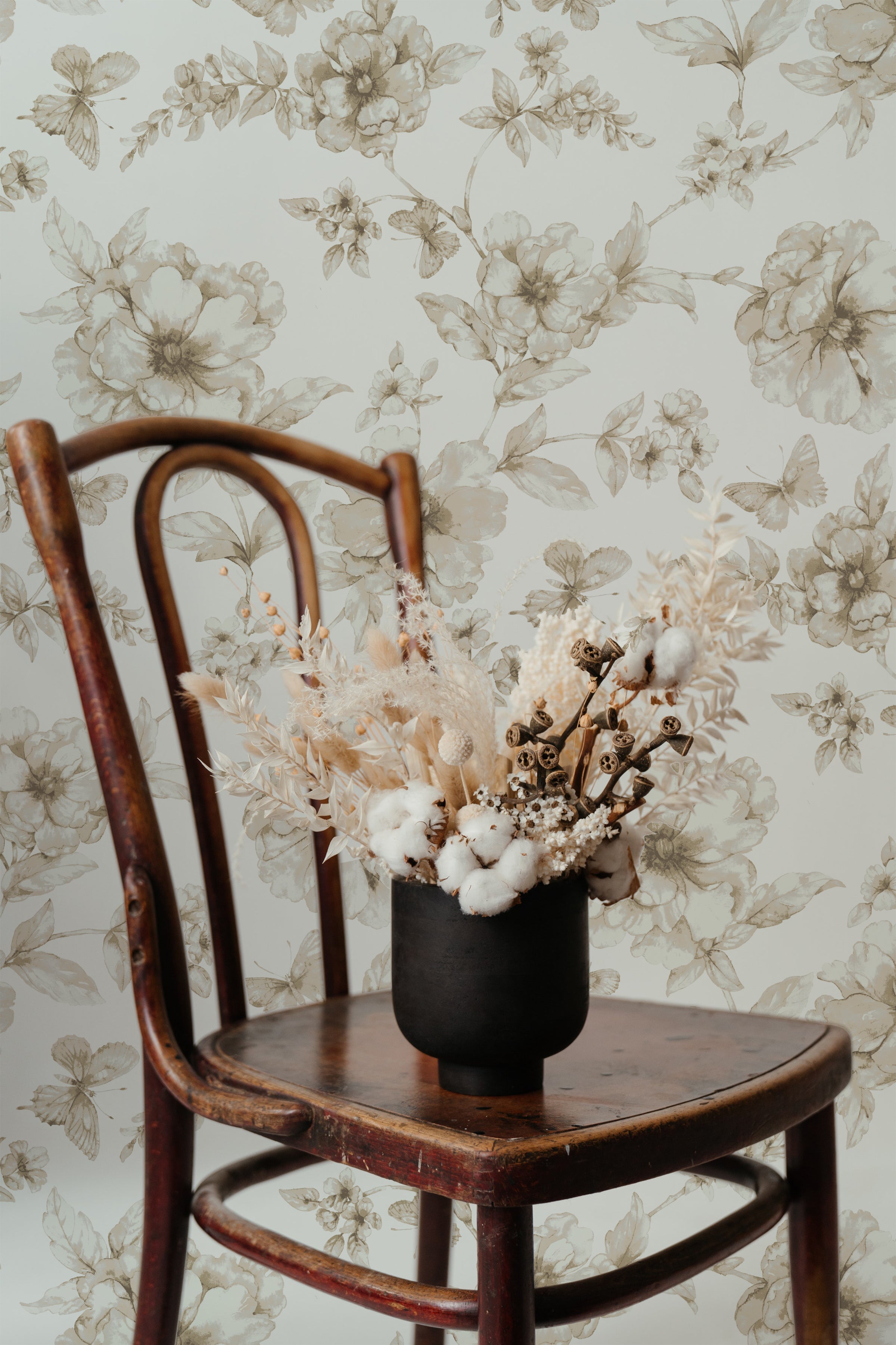An elegant vintage wooden chair in front of a wall adorned with the Sepia Trellis Wallpaper featuring intricate floral designs in sepia tones. A rustic vase with dried flowers complements the timeless appeal of the wallpaper.