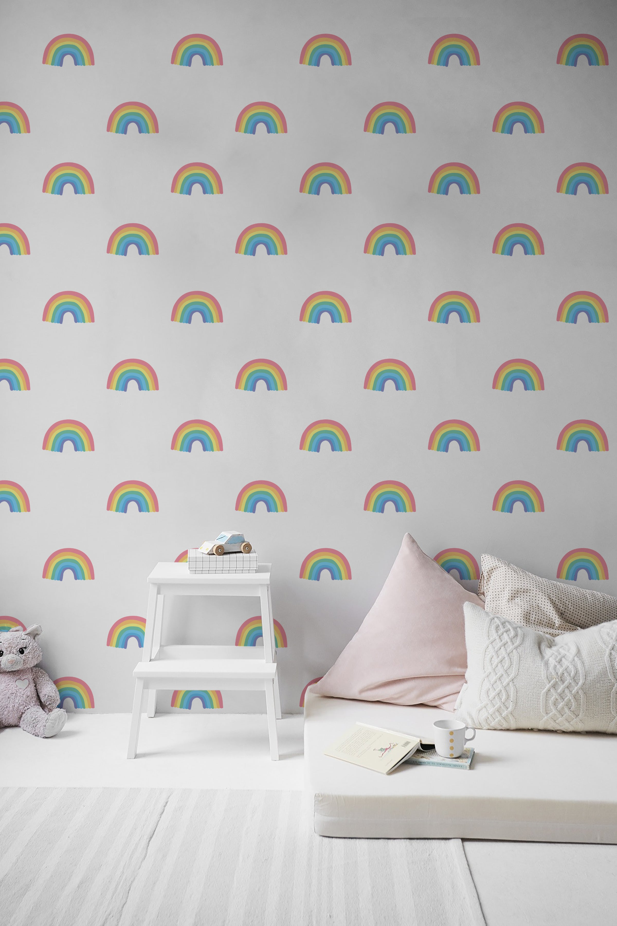 A playful child's room decorated with Chasing Rainbows wallpaper, featuring bright and colorful rainbow patterns on a light gray background. A small step stool, plush toys, and a comfortable couch with cushions complete the cozy setup.