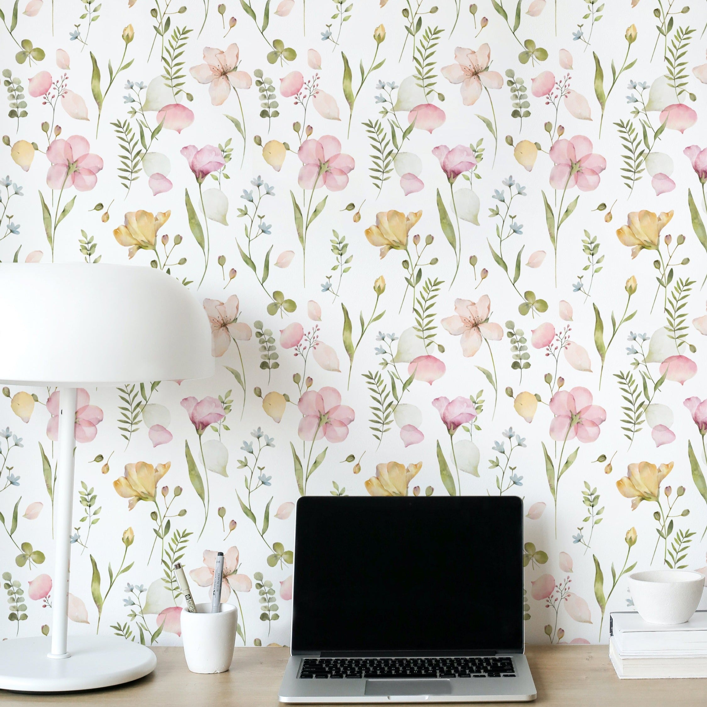 A home office setting decorated with the Serene Floral Wallpaper, providing a vibrant backdrop of soft pink and yellow flowers and green leaves. The desk area is neatly arranged with a laptop, white lamp, and office supplies, creating a peaceful and inspiring work environment.