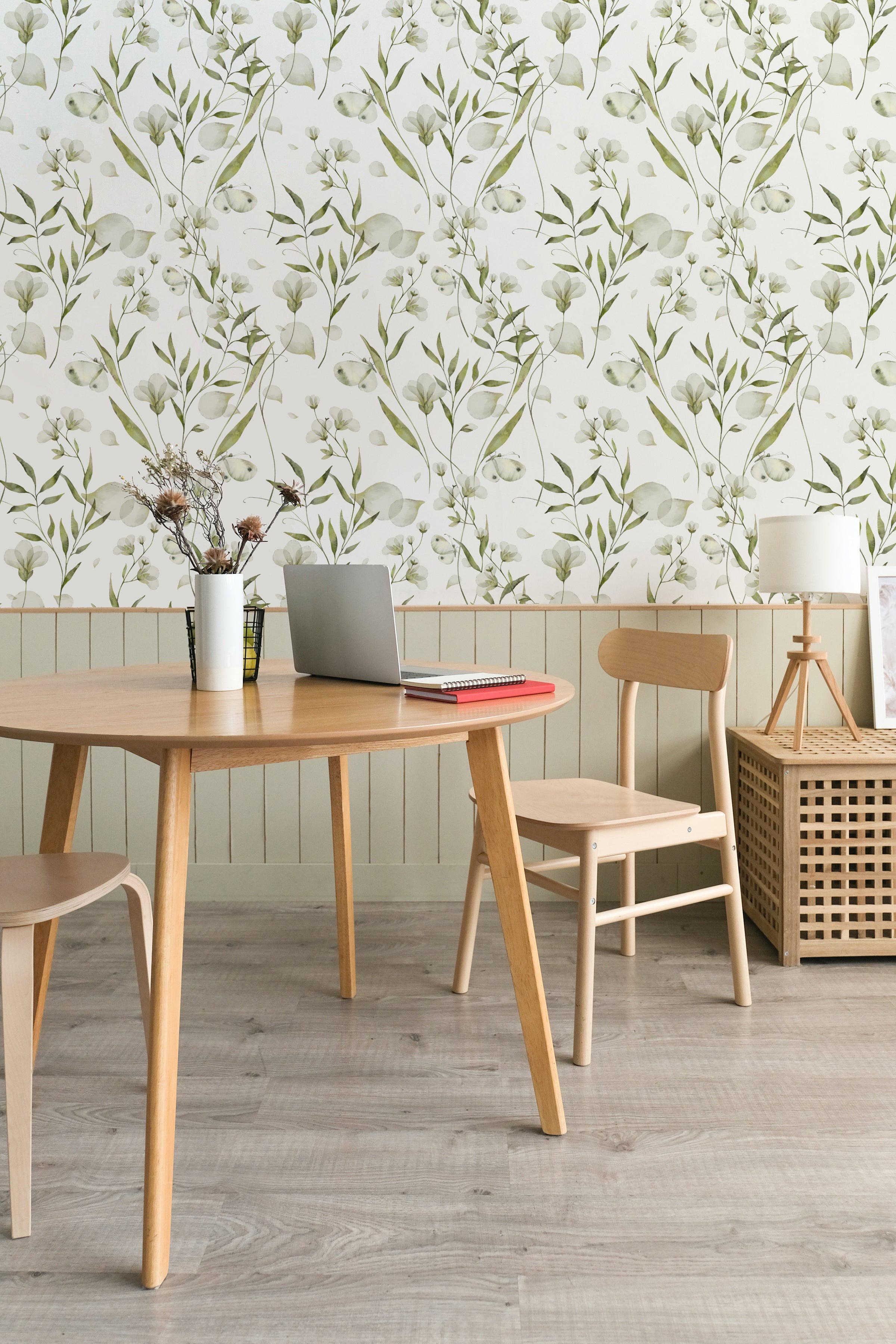 An elegant work area with the 'Botanical Bliss Wallpaper' gracing the wall, featuring a sophisticated pattern of soft green botanicals and white blooms. The natural theme is complemented by a wooden table, chairs, decorative vases with dried plants, and a laptop, creating a harmonious and productive environment