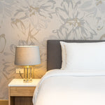 A chic bedroom decorated with the Modernist Meadows Wallpaper featuring oversized floral motifs in soft gray on a beige background. The wallpaper provides a tranquil and stylish backdrop to the modern bed and bedside lamp, creating a refined and inviting atmosphere