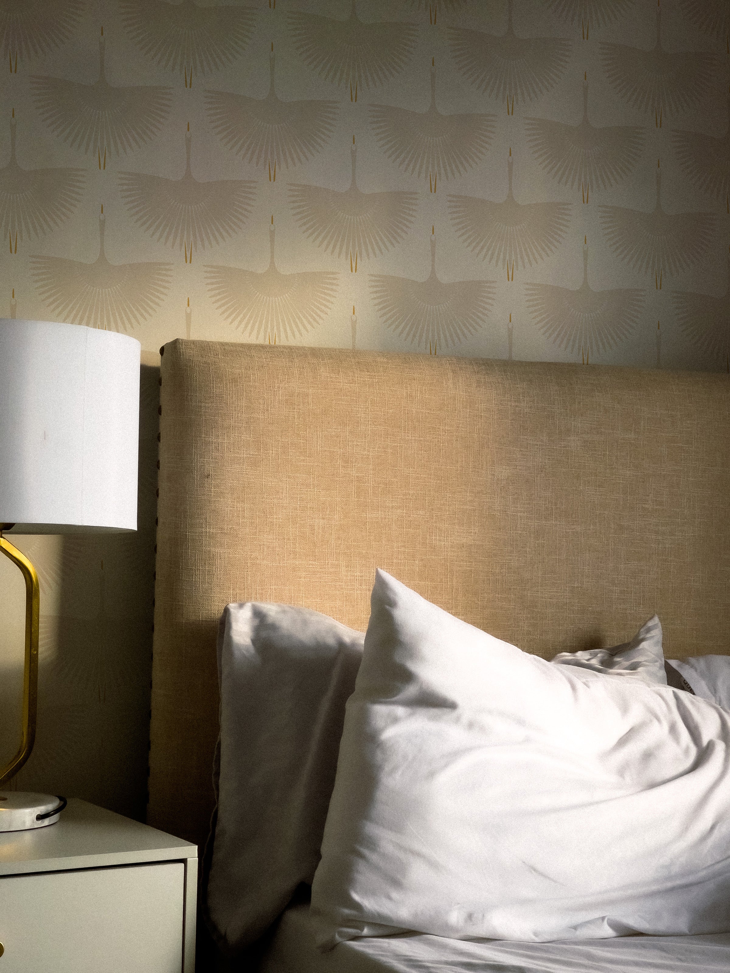 A bedroom scene illuminated by soft ambient lighting that highlights the "Flying Swans Wallpaper". This elegant wallpaper envelops the room where a bed with a simple headboard and white linen is present, creating a restful and stylish sleeping environment.