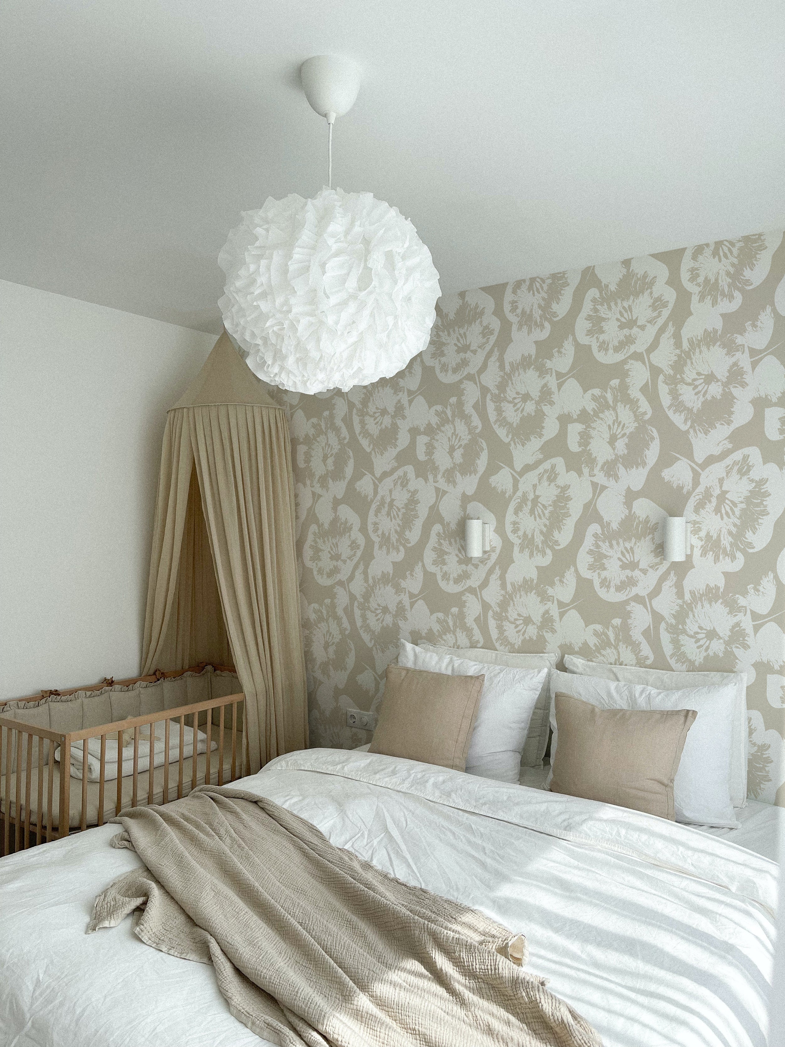 A serene bedroom showcasing the Stamped Floral Wallpaper, where large stylized floral prints in beige provide a soft, romantic touch. The room is furnished with a bed and a crib, unified by the wallpaper's calming presence and a chic white flower-shaped ceiling lamp