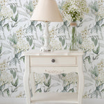An elegant room corner featuring a white vintage table with a drawer, topped with a white lamp with a beige shade and a clear glass vase filled with white flowers. The background is adorned with Heavenly Hydrangea wallpaper, showcasing a pattern of white hydrangea blooms amid soft green and gray foliage.