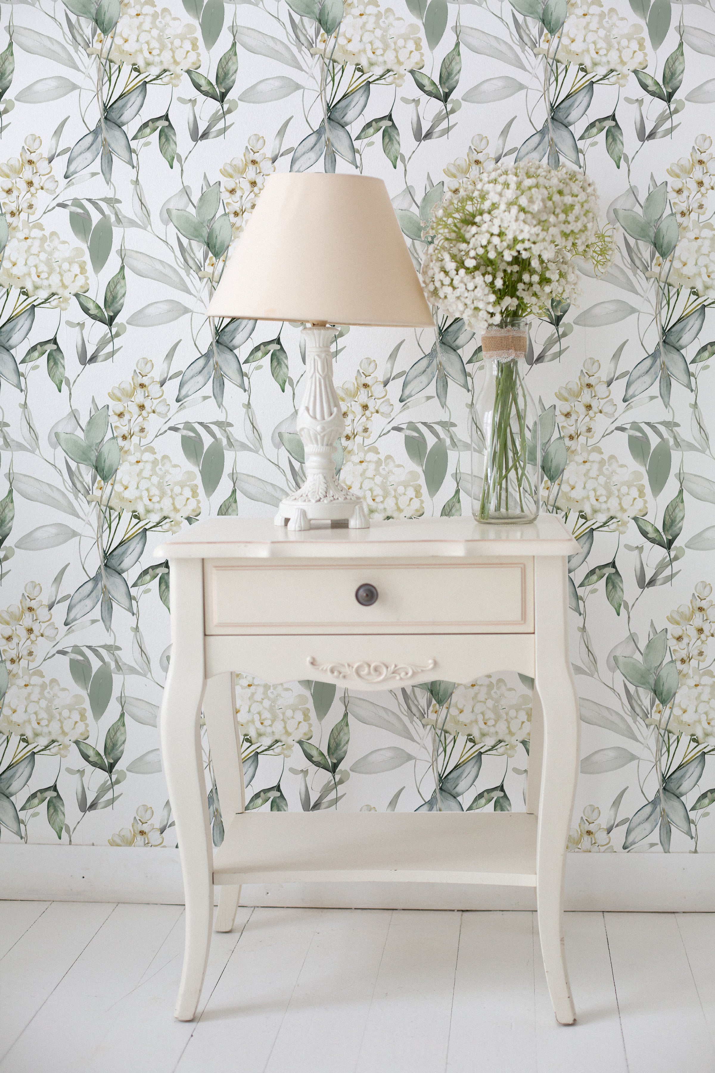 An elegant room corner featuring a white vintage table with a drawer, topped with a white lamp with a beige shade and a clear glass vase filled with white flowers. The background is adorned with Heavenly Hydrangea wallpaper, showcasing a pattern of white hydrangea blooms amid soft green and gray foliage.