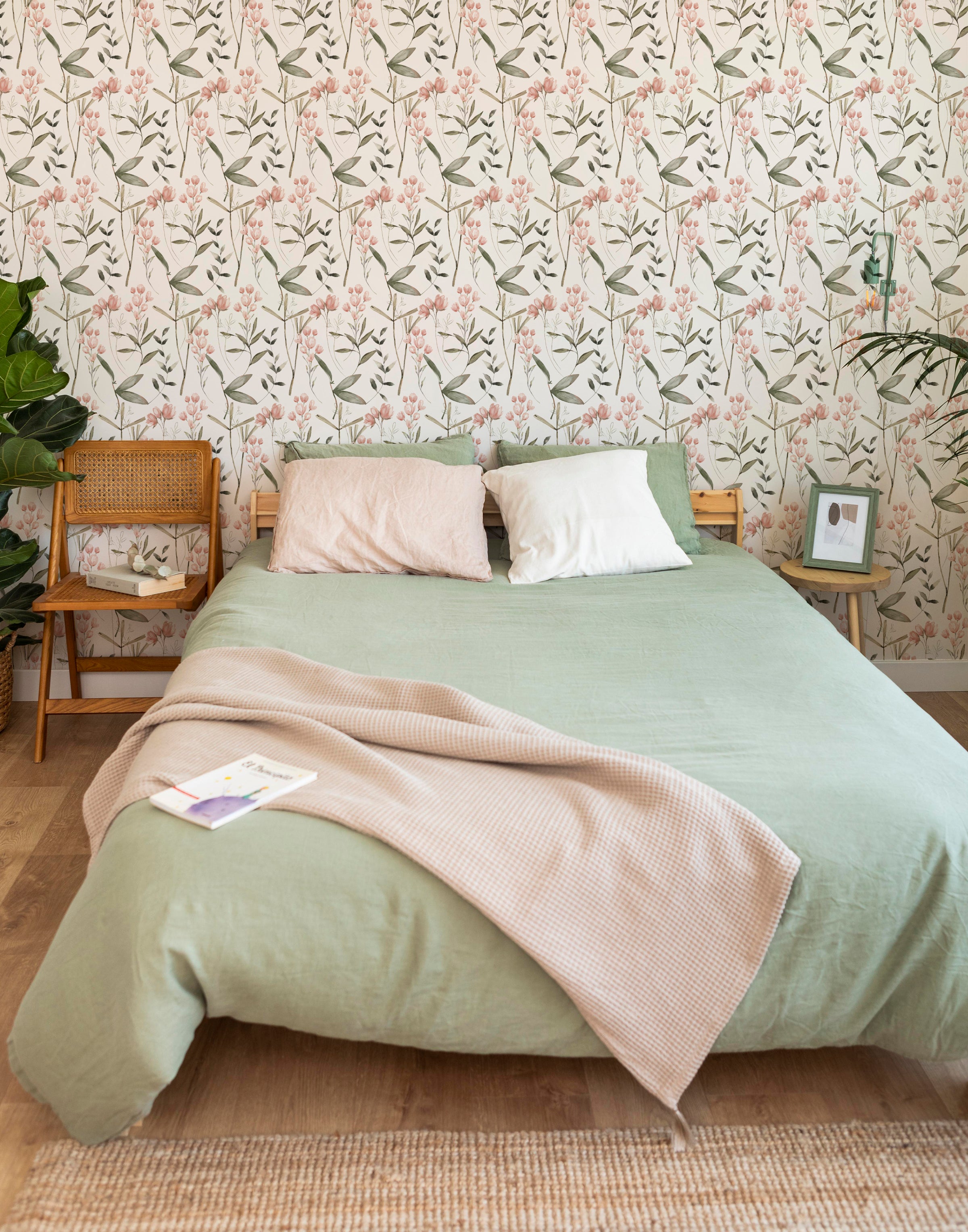 A cozy bedroom featuring the Springtime Sonata Wallpaper, adorned with a delicate pattern of pale pink tulips and lush green leaves against a white background. The elegant floral design complements the earthy tones of the green bedspread and natural wood furniture, creating a tranquil and inviting space.