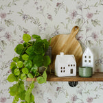 Elegant floral wallpaper with intricate botanical prints of flowers and leaves in muted shades of green and pink, beautifully complemented by home decor items like wooden cutting boards, ceramic houses, and a lush green plant on a rustic wooden shelf
