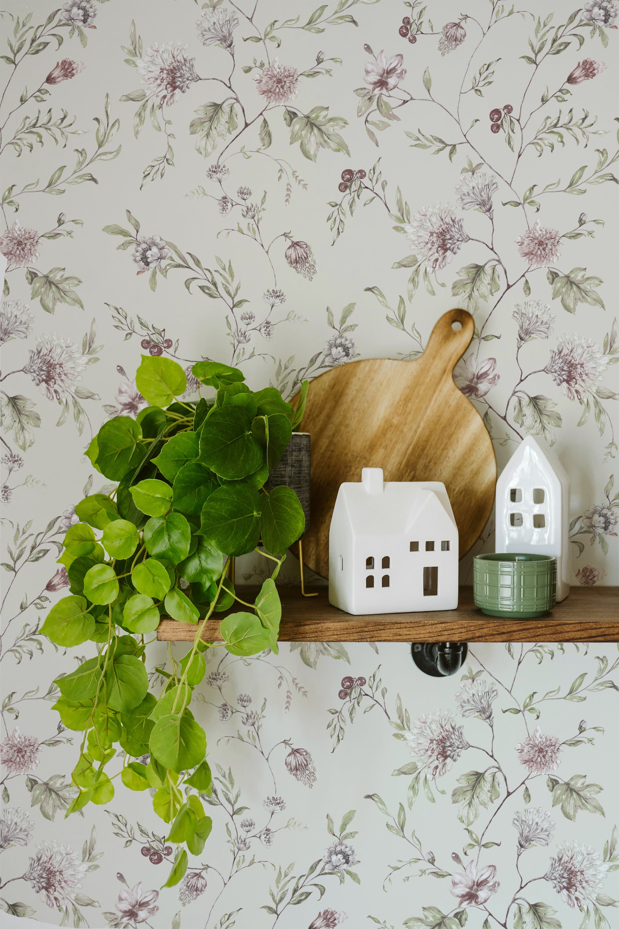 Elegant floral wallpaper with intricate botanical prints of flowers and leaves in muted shades of green and pink, beautifully complemented by home decor items like wooden cutting boards, ceramic houses, and a lush green plant on a rustic wooden shelf