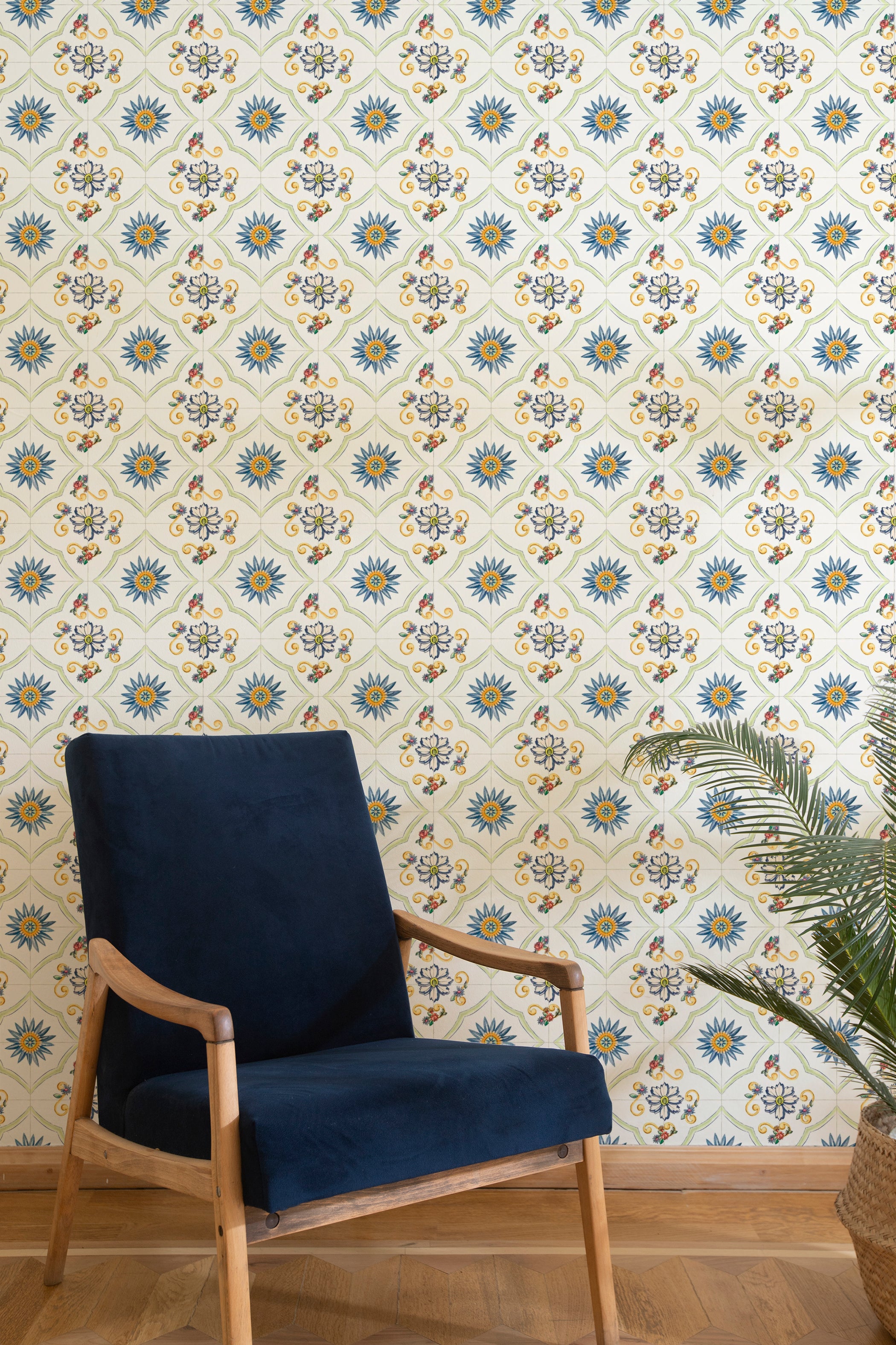 A cozy living room corner featuring a navy blue upholstered wooden chair against a backdrop of Spanish Tile Wallpaper, which displays a repetitive pattern of blue and yellow sunbursts with floral accents. The room has a light wooden floor, and a potted palm adds a touch of greenery.