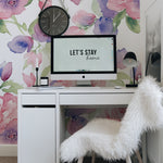 A stylish home office setup featuring Mystic Garden Wallpaper, which displays a lush array of watercolor roses and other blooms in shades of pink, purple, and blue. Above the minimalist white desk, a motivational 'Let's stay home' poster hangs on the floral backdrop, creating a serene and inspiring workspace.