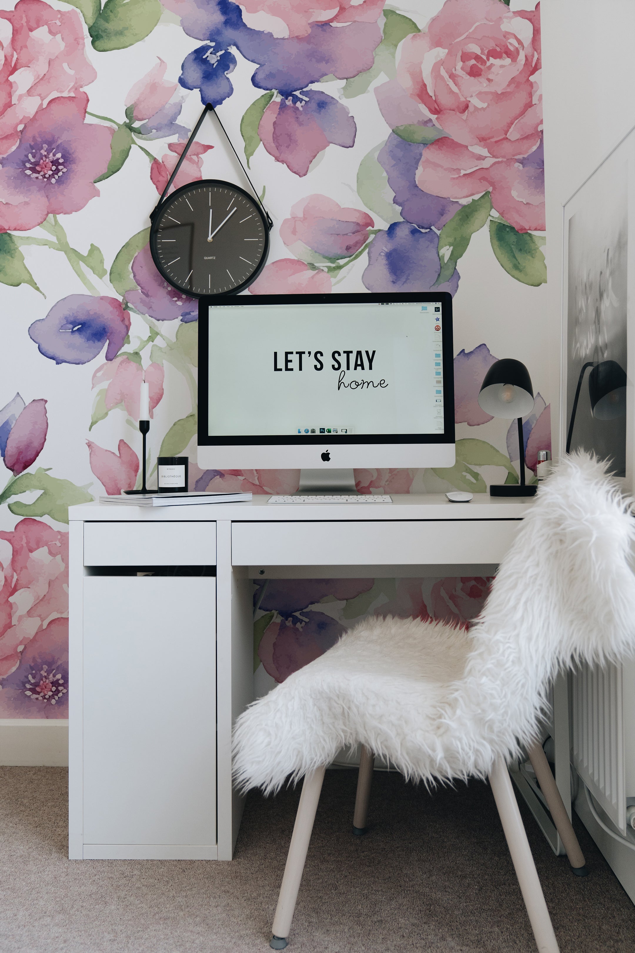 A stylish home office setup featuring Mystic Garden Wallpaper, which displays a lush array of watercolor roses and other blooms in shades of pink, purple, and blue. Above the minimalist white desk, a motivational 'Let's stay home' poster hangs on the floral backdrop, creating a serene and inspiring workspace.