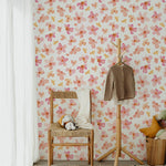A charming nursery room decorated with Peachy Floral Wallpaper, featuring pink and peach watercolor flowers. The room includes a wooden chair with a straw seat, a stuffed animal, and a small children’s coat rack with a sweater. Soft-colored pillows and a round rug complement the gentle tones of the wallpaper.
