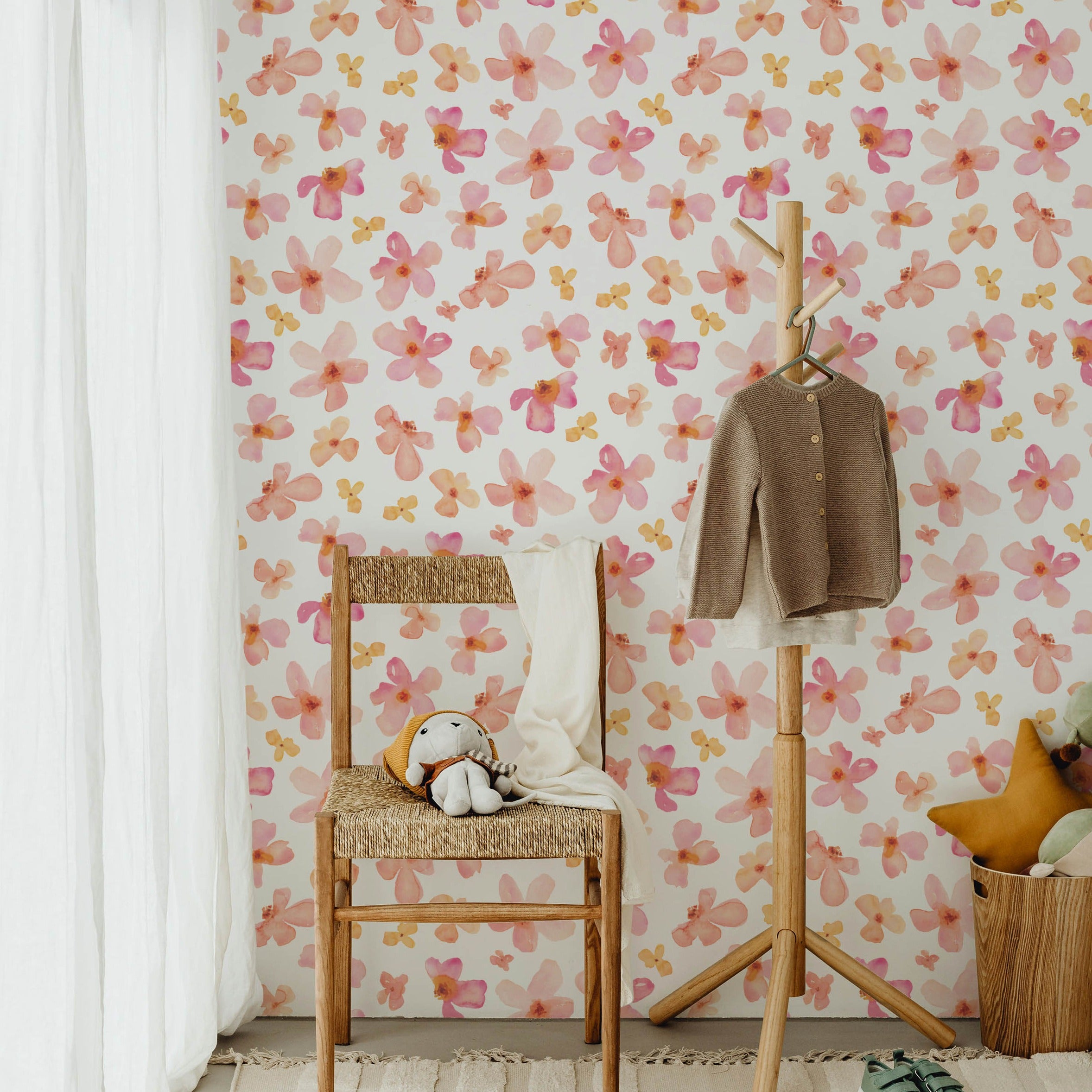 A charming nursery room decorated with Peachy Floral Wallpaper, featuring pink and peach watercolor flowers. The room includes a wooden chair with a straw seat, a stuffed animal, and a small children’s coat rack with a sweater. Soft-colored pillows and a round rug complement the gentle tones of the wallpaper.