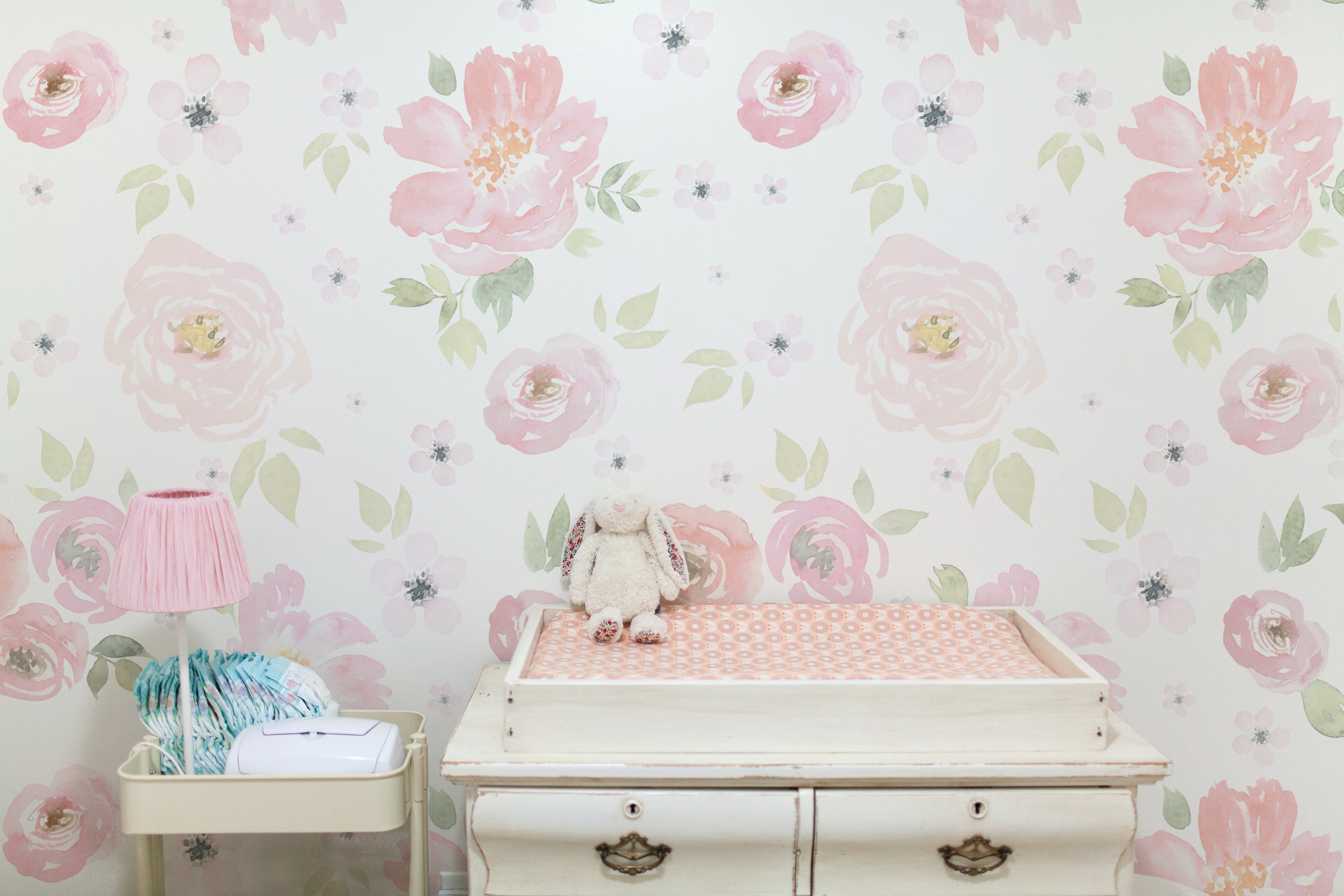 A charming nursery scene with a vintage white changing table adorned with a plush toy bunny, under a wall covered in 'Gentle Blossom Wallpaper' featuring large watercolor flowers in soft pink and green tones, creating a soothing and whimsical atmosphere.