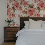 A bedroom interior showcasing one wall adorned with 'Grace Floral Wallpaper', which features large pink flowers and soft green leaves. The room includes a wooden bed with white bedding, a small wooden nightstand with a white lamp and a small pine tree decoration, creating a cozy and inviting atmosphere