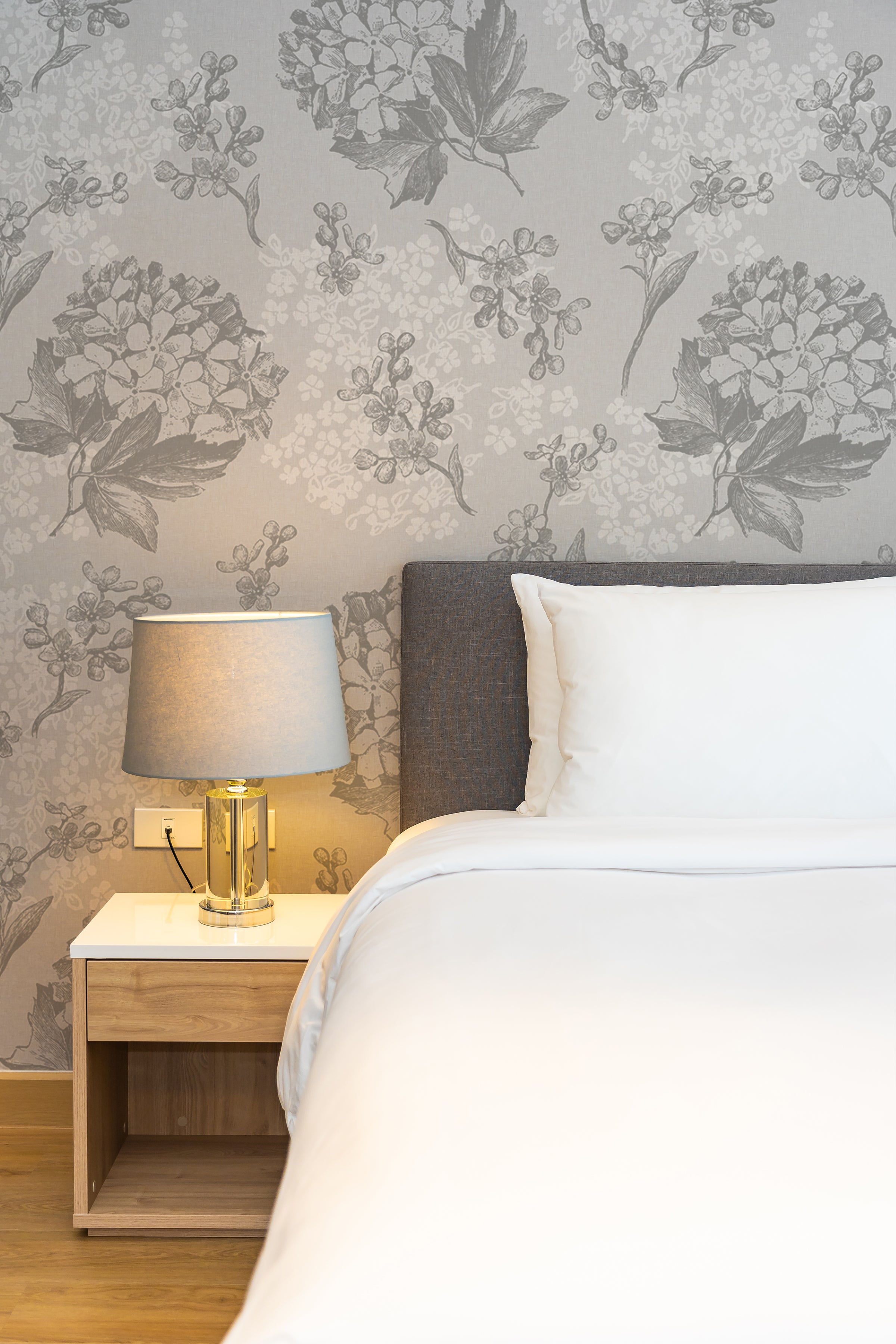 Chic bedroom featuring Modern Hydrangea Wallpaper with large grey floral patterns of hydrangeas on a muted background. The design complements a contemporary bedside setting with a sleek table lamp and minimalist furnishings, creating a tranquil and stylish ambiance.
