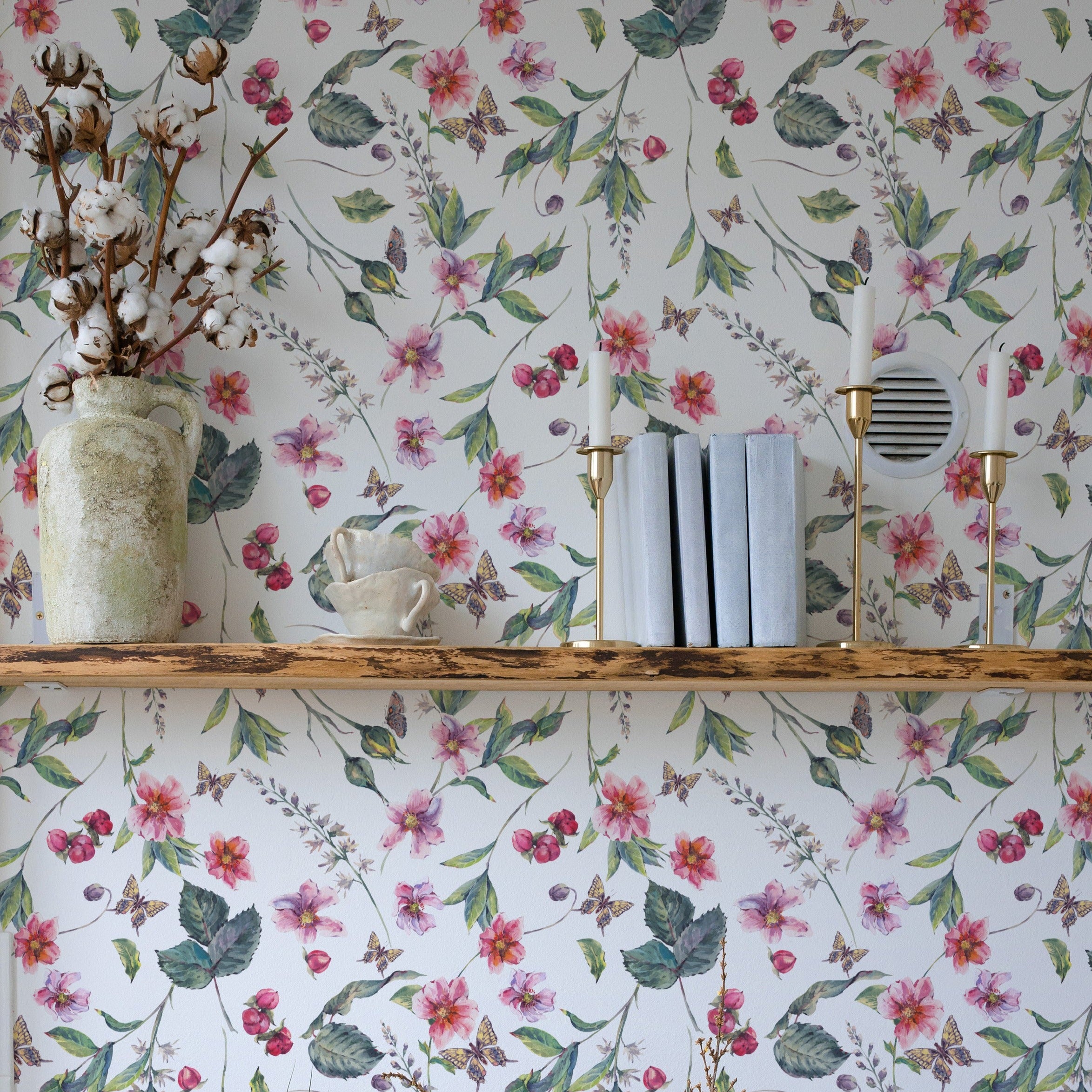 Charming display on a wooden shelf against Graceful Garden Wallpaper, which is adorned with pink flowers, green leaves, and butterflies. Decor includes a rustic vase with dried cotton stems and small books.
