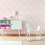 a child's room with "Pretty Petals Wallpaper - 12.5" on the wall. The wallpaper is adorned with a delicate floral pattern in shades of pink and beige, creating a soft, pastel backdrop. The room features a white modern rocking horse, a sideboard with colorful baskets, and a lamp with pink legs, conveying a playful yet stylish nursery design.