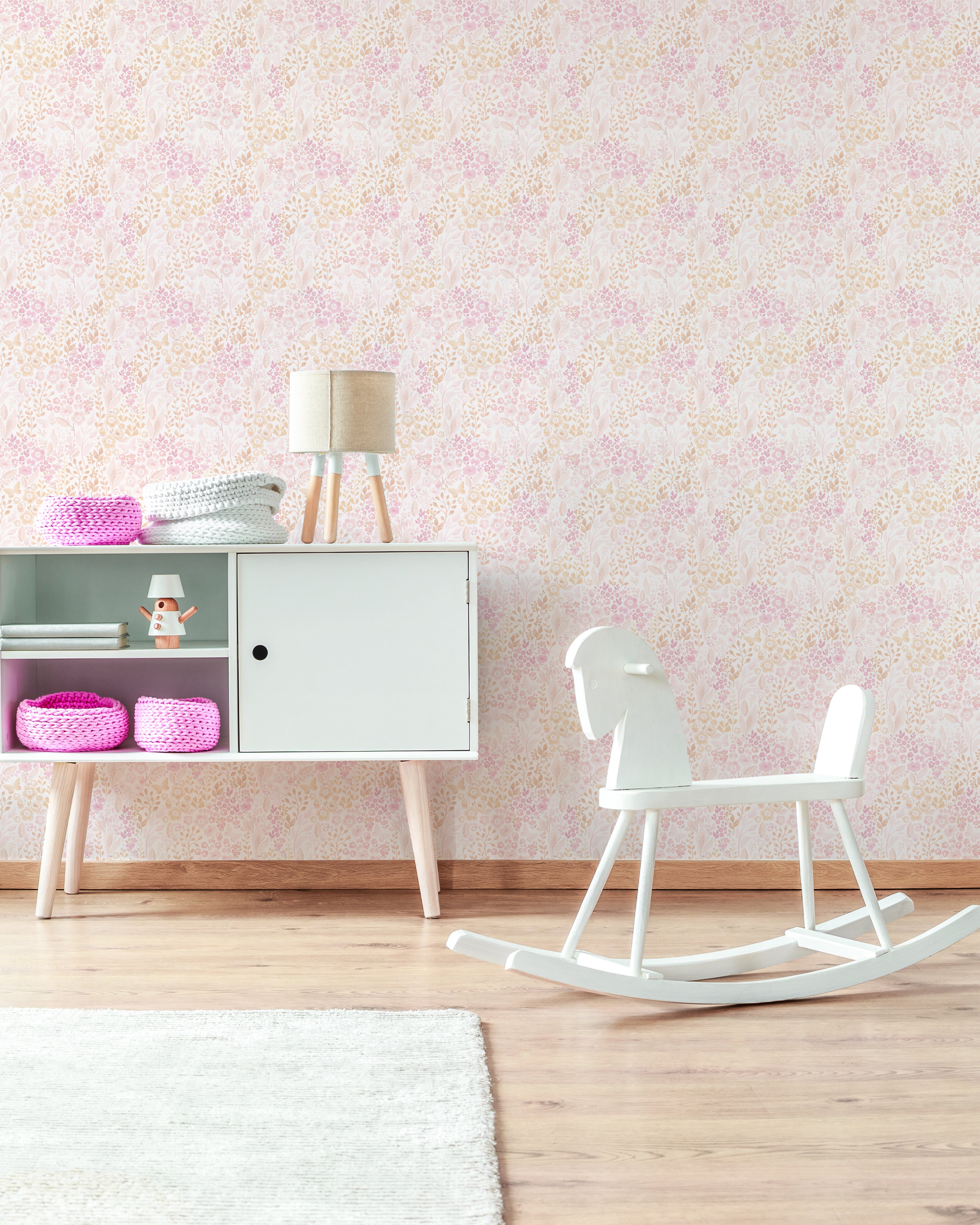 a child's room with "Pretty Petals Wallpaper - 12.5" on the wall. The wallpaper is adorned with a delicate floral pattern in shades of pink and beige, creating a soft, pastel backdrop. The room features a white modern rocking horse, a sideboard with colorful baskets, and a lamp with pink legs, conveying a playful yet stylish nursery design.