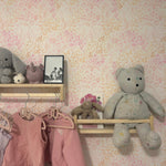 a nursery setting with "Pretty Petals Wallpaper - 12.5"," as the focal point. The wall is decorated with the pastel floral pattern, complemented by a display of children's clothing, plush toys, and a framed photograph, enhancing the room's sweet and cozy ambiance.
