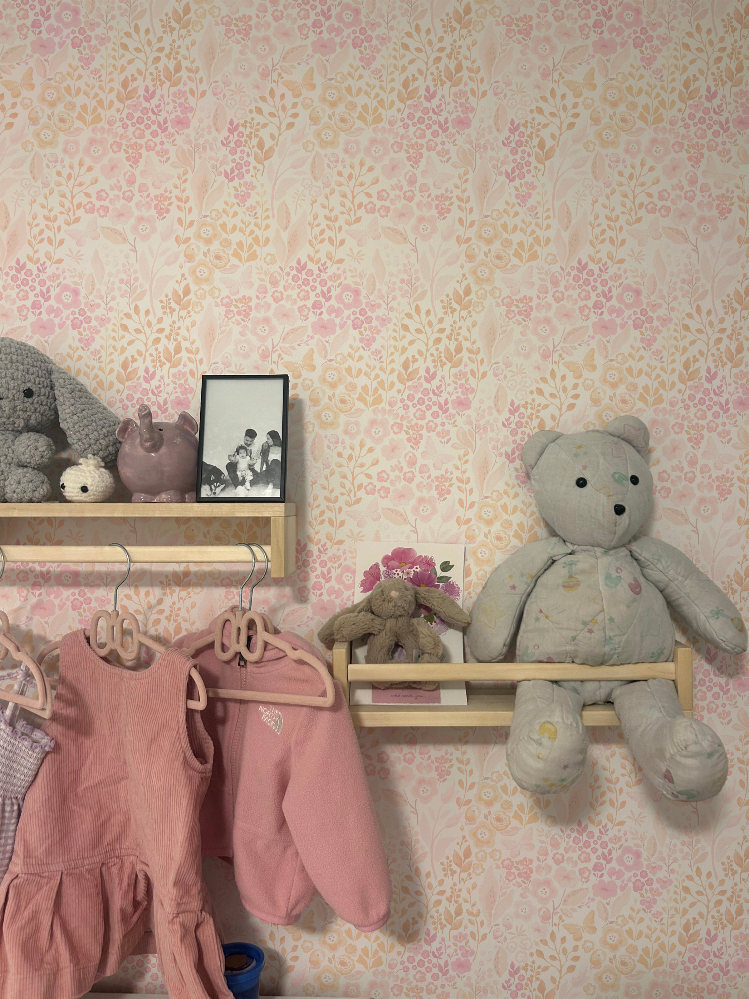 a nursery setting with "Pretty Petals Wallpaper - 12.5"," as the focal point. The wall is decorated with the pastel floral pattern, complemented by a display of children's clothing, plush toys, and a framed photograph, enhancing the room's sweet and cozy ambiance.
