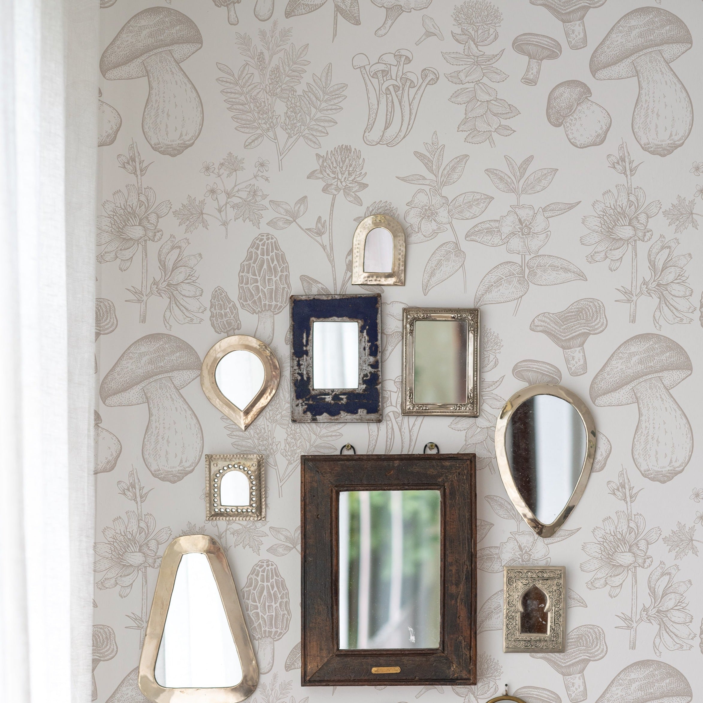 An eclectic collection of vintage mirrors in various shapes and sizes adorns a wall covered with 'Mushroom Garden Wallpaper'. The wallpaper features a detailed botanical print with various species of mushrooms and plants in shades of beige, providing a whimsical backdrop.